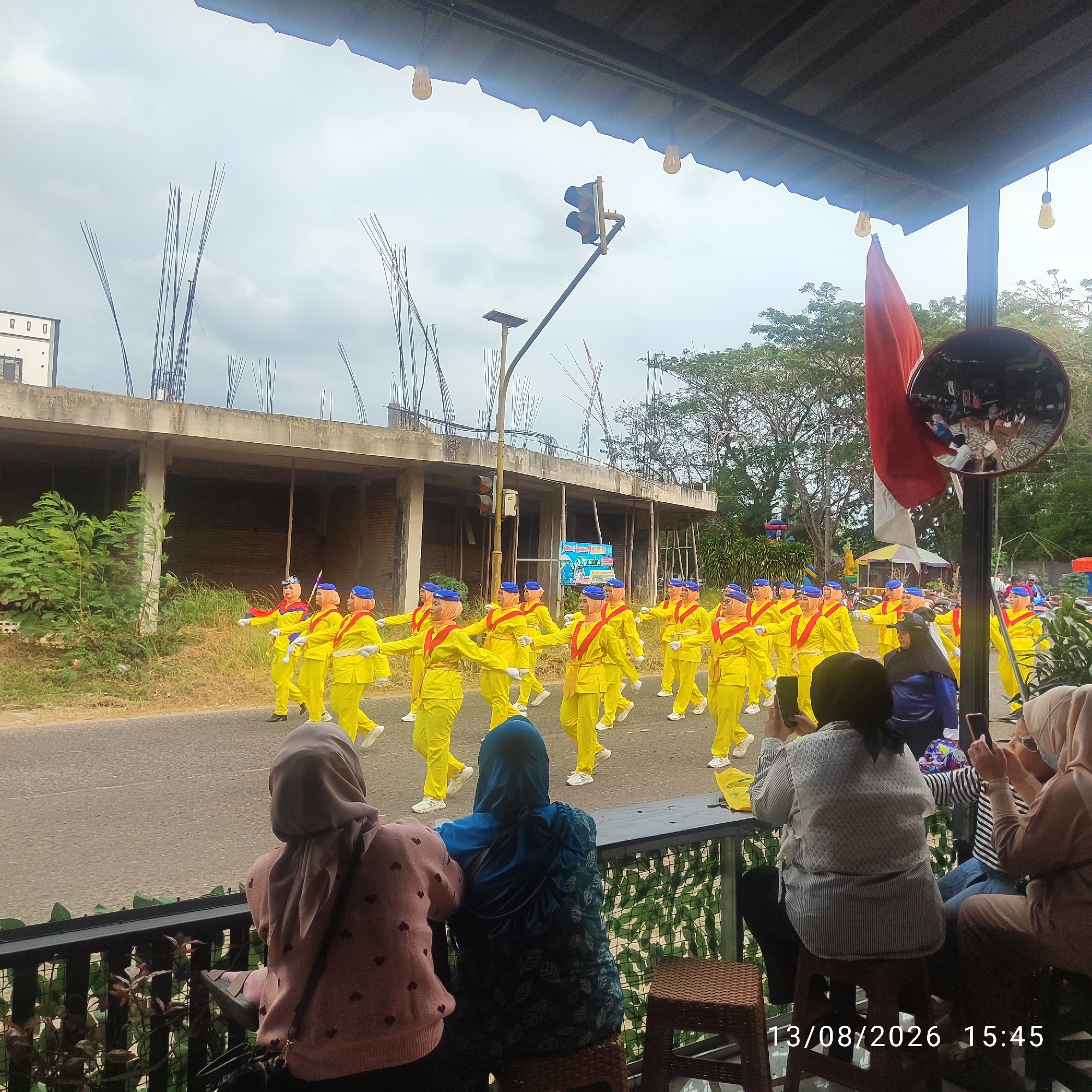 In Buding, Indonesia, a group of individuals in yellow uniforms with blue headscarves and red sashes are marching in formation on a paved road. They are in the middle of a parade or procession. In the foreground, three women are seated, facing away from the camera, watching the parade. One woman in a pink sweater with heart patterns is on the left, another in a blue patterned outfit is in the middle, and a woman in a beige hijab is on the right. All three appear to be holding or using their phones. A black fence separates the seated observers from a railing and greenery. In the background, a building under construction with exposed rebar stands beside the road. A traffic light is visible on a pole, and a red and white flag is displayed prominently. The sky is overcast. The date and time "13/08/2026 15:45" are superimposed on the lower right of the image.
