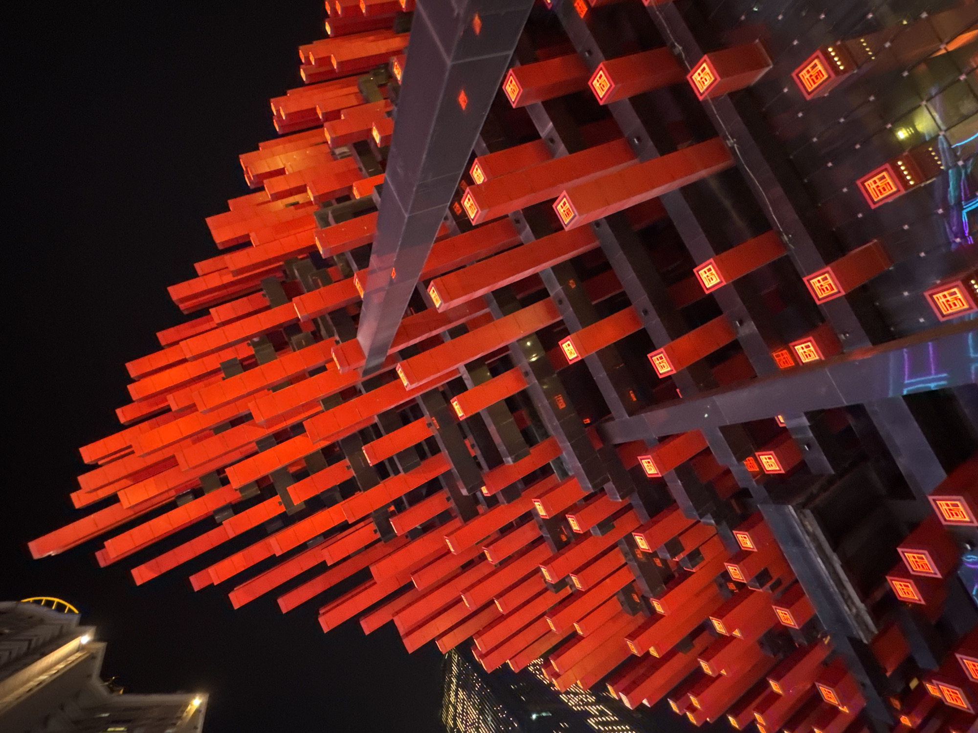 A low-angle shot shows a massive, illuminated public art installation in the Yuzhong District, China. The installation consists of a grid of gray support beams, with hundreds of horizontal, rectangular red bars protruding from them. Each red bar has a square, illuminated design on its end. The background is a dark night sky, with buildings visible at the bottom of the frame, lit by artificial lights. The overall scene is a brightly lit, geometric display against a dark backdrop in an urban setting.