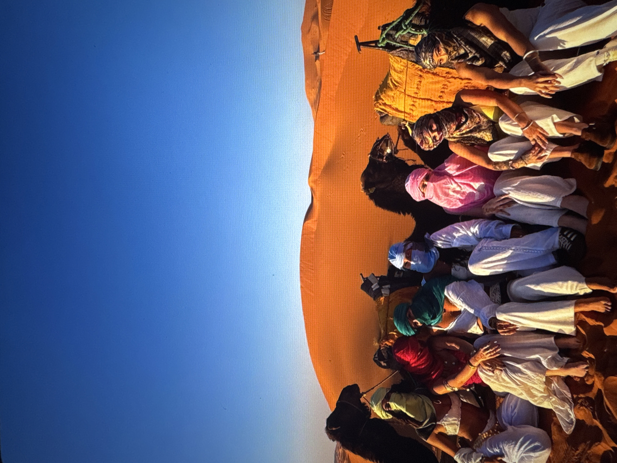 In a desert landscape under a clear blue sky, a group of nine individuals are seated on the sand, with camels positioned behind them. The people are adorned with colorful headscarves and traditional desert attire, with their faces partially or fully covered. They are arranged in two rows, with the front row consisting of five individuals and the back row of four. In the background, sand dunes stretch across the horizon, illuminated by the bright sunlight. The scene depicts a travel experience in a desert environment. The image does not contain any elements or context that would associate it with the city of Tallinn, Estonia.