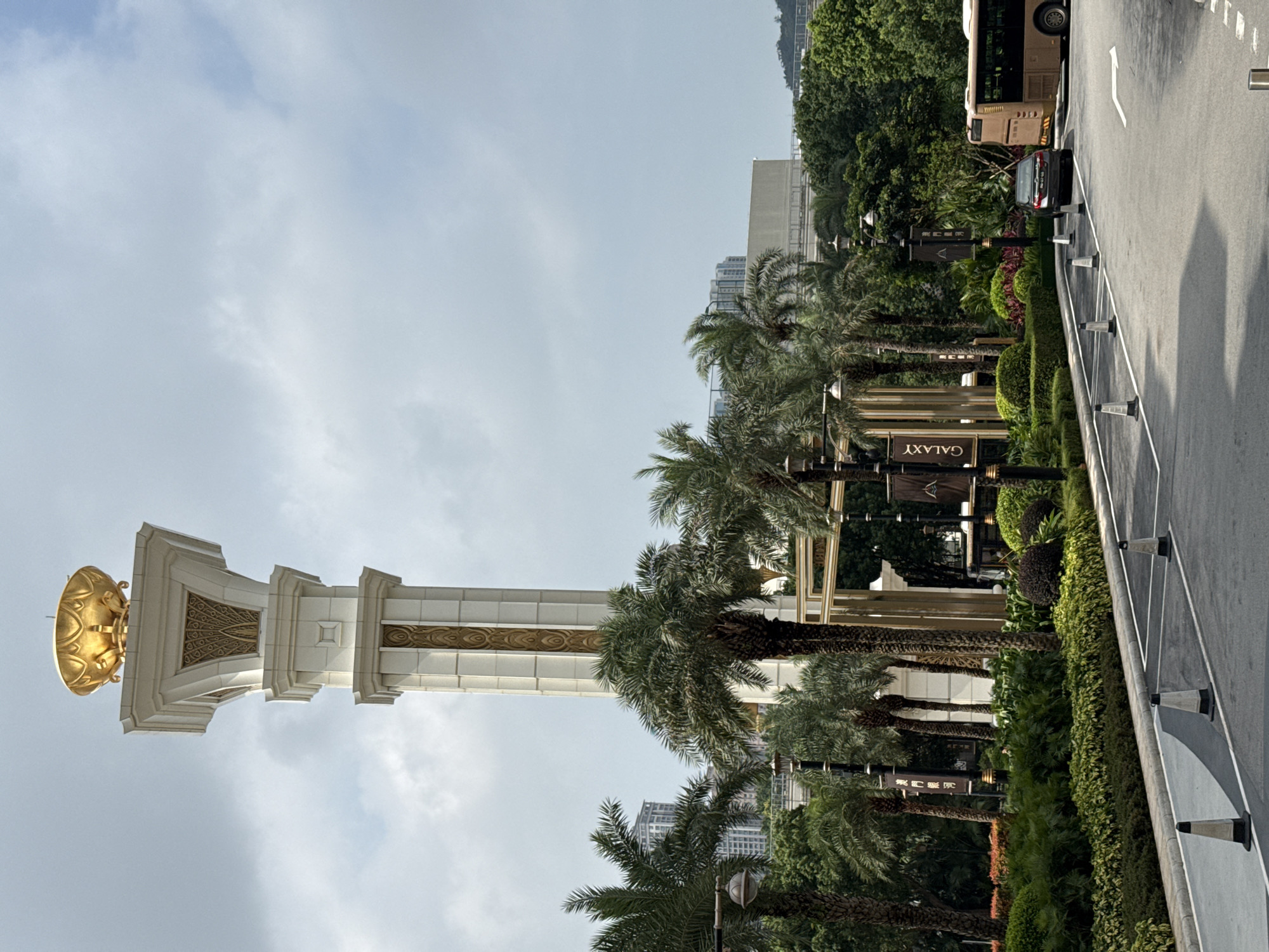 A tall, ornately decorated white and gold column with a large golden sphere on top stands in front of palm trees, signage for the Galaxy Hotel, and other greenery. A street runs along the right side, with traffic control devices, a bus, and a car visible. The sky is overcast. The location is Unknown, Macao.