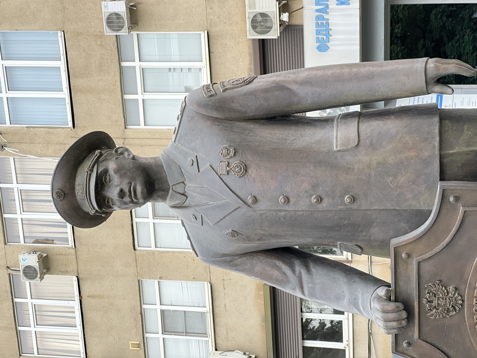 The image depicts a bronze statue of a uniformed man in front of a building in Krasnodar, Russia. The statue stands to the right, facing toward the left of the frame. The man wears a military-style uniform with a hat and several medals and insignias on the chest and shoulders. His face is detailed and appears to be looking slightly down. He holds onto a decorated shield or plaque with his right hand.
Behind the statue, there is a light brown building with rows of rectangular windows. Some of the windows have white frames and are filled with blinds. Air conditioning units are mounted on the building's exterior, near the windows. The building also displays blue lettering in Cyrillic script, indicating "ФЕДЕРАЛ." The background shows signs of greenery, suggesting trees or plants.