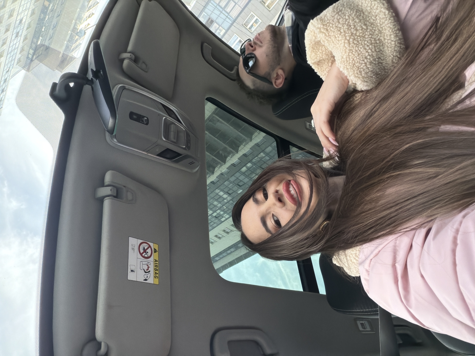 The image shows the interior of a car, specifically the front seating area. A woman with long brown hair and a light pink jacket is visible, angled towards the camera from what appears to be the passenger seat. A man wearing sunglasses is also partially visible near the sunroof. The car's interior includes a rearview mirror, sun visors (one with an airbag warning label), and a sunroof. Through the sunroof, there are buildings and a cloudy sky, potentially locating the scene in an urban environment, possibly Saint Petersburg, Russia, based on building styles.