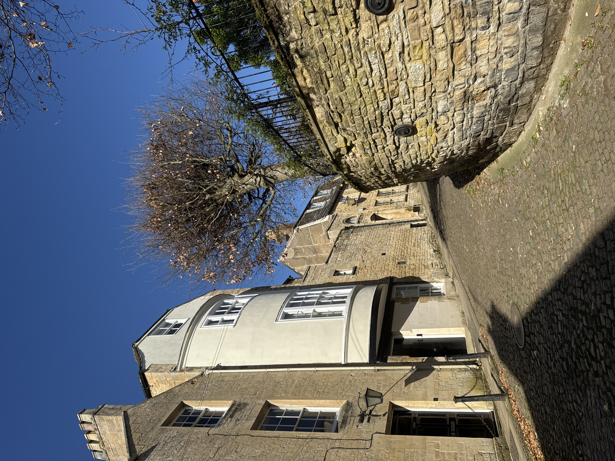 An upward-angled view showcases a narrow streetscape in South Kesteven, UK, dominated by architectural elements and natural features. The buildings are constructed of stone and brick, featuring windows and varying rooflines. A stone wall curves along the right edge of the image, topped by a black metal fence with greenery. A tree with bare branches and some brown leaves extends across the blue sky. The street is paved with cobblestones.