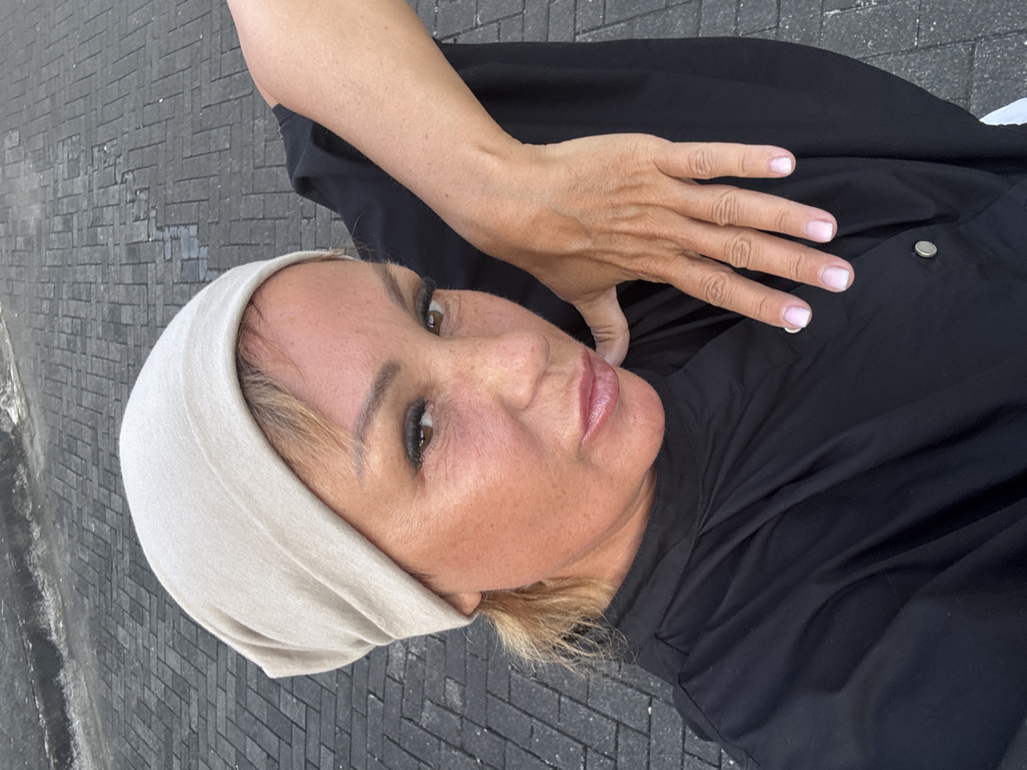 A portrait-style image captures a woman from a high angle, with the image rotated 90 degrees clockwise. She presents a calm expression with a subtle, closed-mouth smile. Her face is framed by a light beige or off-white head covering, beneath which light brown or blonde hair is visible. She has brown eyes with subtle makeup and her lips are adorned with light pink lip gloss.
She is wearing a black garment, possibly a shirt or dress, which has a visible silver snap button on its front. Her right arm and hand are prominent, with her hand gently resting on the black fabric. Her fingernails are neatly manicured with a natural or light pink polish.
The background consists of a dark gray paved surface, likely ground, made of rectangular bricks laid in a herringbone pattern. The lighting appears soft and even, suggesting an overcast day or a shaded outdoor environment. No text is visible in the image. The setting is outdoors, in what appears to be an urban or paved area, within the context of Kuta Selatan, Indonesia.
