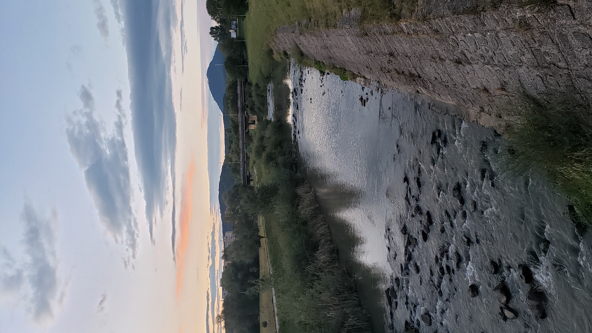 The image depicts a river flowing through a park-like area with a bridge in the background. The sky is partly cloudy with a hint of pinkish hues, suggesting either sunrise or sunset. On the left bank of the river, there is a grassy area with trees and some buildings in the distance. A paved path runs along the riverbank, and a lone figure is seen walking on it. The right bank of the river is lined with a stone embankment. The river itself has a rocky bottom in the foreground, with water flowing over the rocks. In the far background, a prominent hill with a pointed summit is visible. The scene appears to be a tranquil urban or suburban landscape, possibly the area around the Sekčov district in Prešov, Slovakia, given the characteristics of the landscape including the river, park, bridge, and surrounding hills.