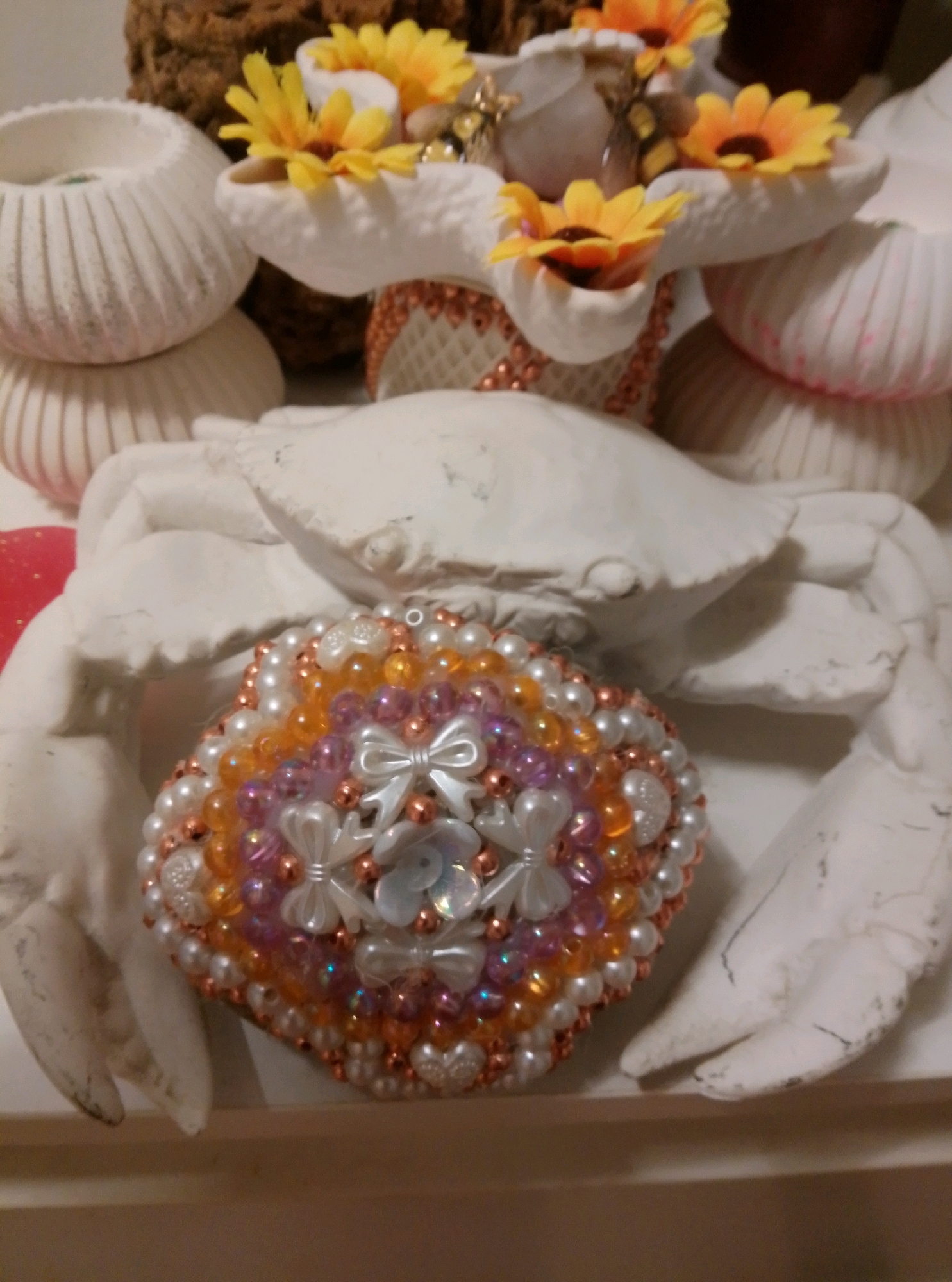 A close-up, eye-level indoor shot displays a decorative white plaster crab sculpture. The crab's body is adorned with a heart-shaped, beaded embellishment featuring pearls, orange and pink beads, and white bows and sequins. The crab sculpture is positioned in front of white ribbed decorative bowls and yellow artificial flowers. The background includes a textured brown object, possibly a piece of driftwood or cork. The lighting is dim and focused on the central subjects. The scene is part of a personal collection or display, likely in a home setting in Wedel, Germany.