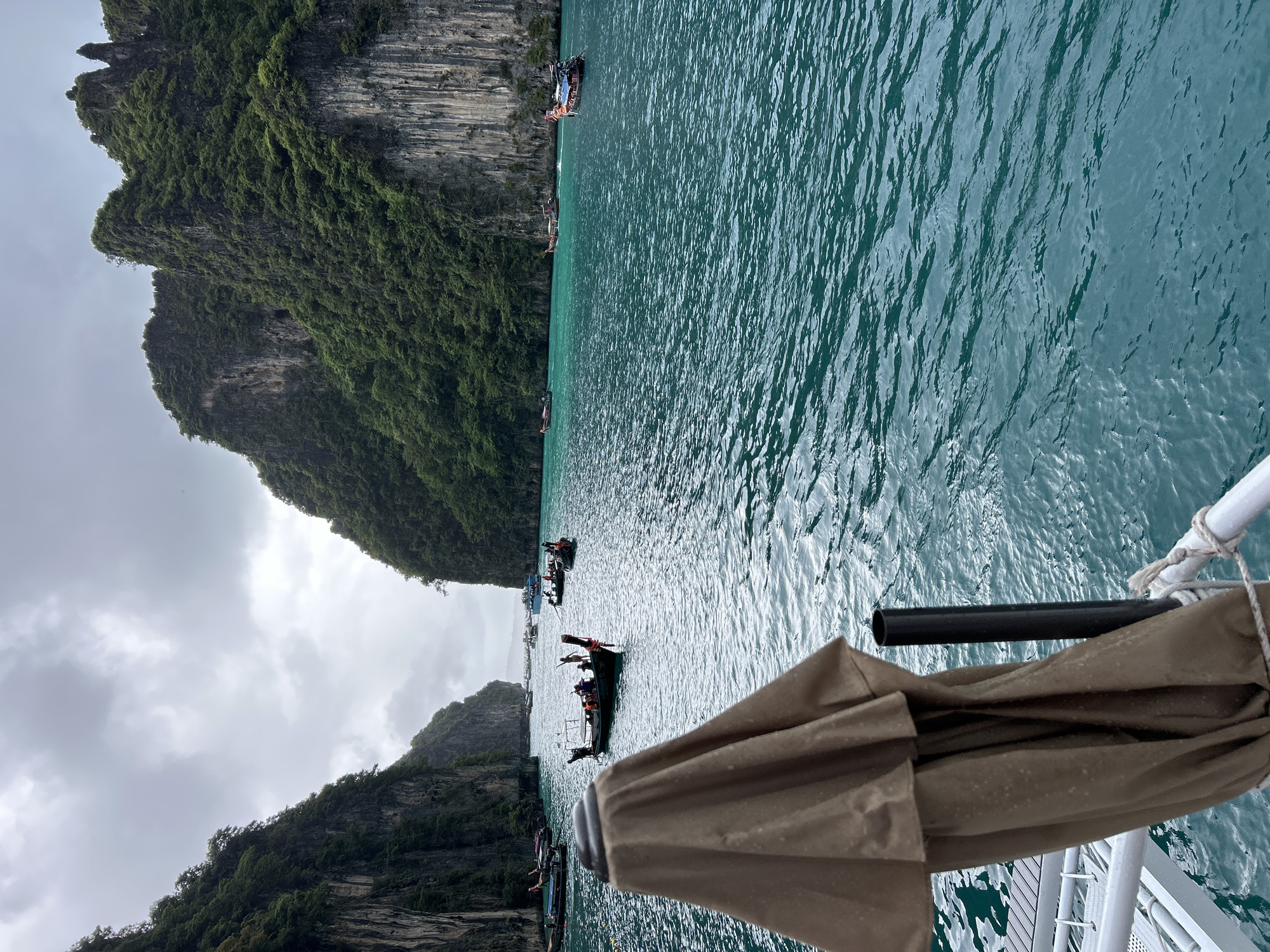 This image depicts a view of Maya Bay in Ao Nang, Thailand, on an overcast day. The foreground features a tan-colored umbrella, partially obscuring a white railing and a black pole. Beyond the railing, the turquoise waters of the bay are visible, with several longtail boats dotting the surface. Some boats have passengers, indicated by the presence of multiple figures. In the mid-ground and background, towering limestone cliffs covered in lush green vegetation rise dramatically from the water. The sky is heavy with clouds, contributing to a diffused light across the scene. Several more boats are scattered further out in the bay, suggesting a moderate level of tourist activity. The overall impression is one of natural beauty within a popular tourist destination.