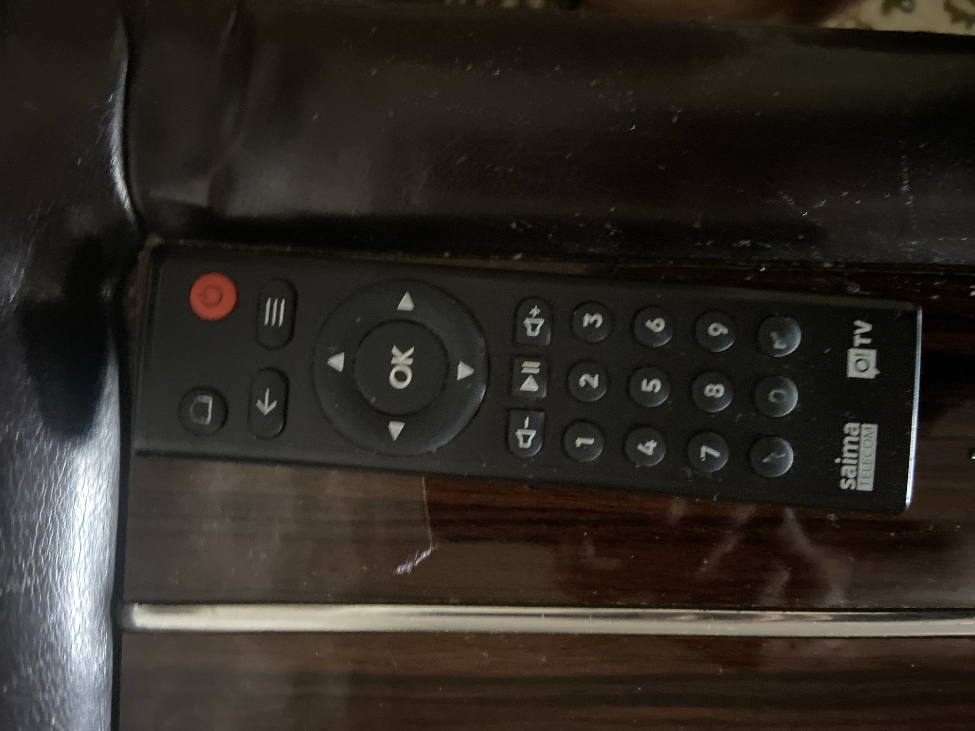 The image shows a black TV remote control with white buttons and text. The remote has numbered buttons from 0 to 9, as well as navigation buttons and a red power button. The brand name "saima TELECOM" and "Oi TV" are visible at the bottom of the remote. The remote is resting on a dark, wooden surface that appears to be a table or furniture. The lighting in the image is dim, and the surface has some dust and scratches. The background is out of focus, but it appears to be a dark, possibly upholstered surface. There is no indication of the city of Kojomkul, Kyrgyzstan, in the image itself or in the provided crops.