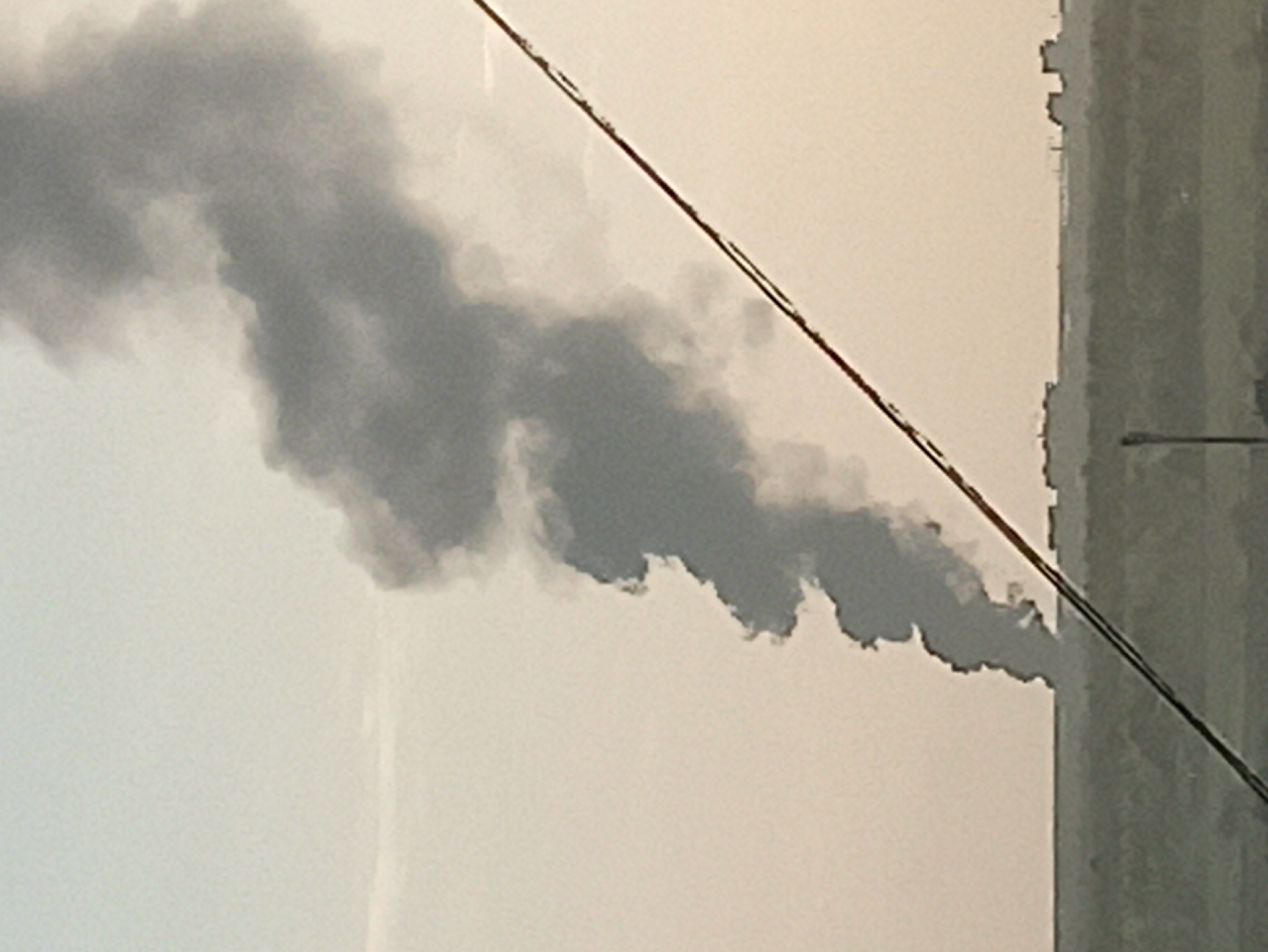 The image depicts a column of dark grey smoke emerging from a structure, possibly an industrial building, in what appears to be an urban or semi-urban environment. The sky is overcast. An electrical wire bisects the frame diagonally. Part of the vertical structure from which the smoke is emitting is visible on the right of the frame. Without more information, a precise location within Кондратово, Russia cannot be determined. There are no people visible.