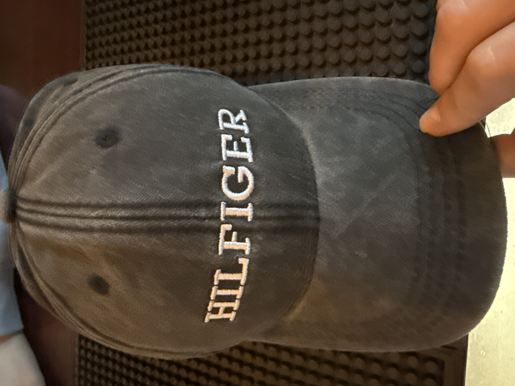 A gray "HILFIGER" baseball cap is lying on a dark brown textured surface. A hand is holding the right edge of the bill.