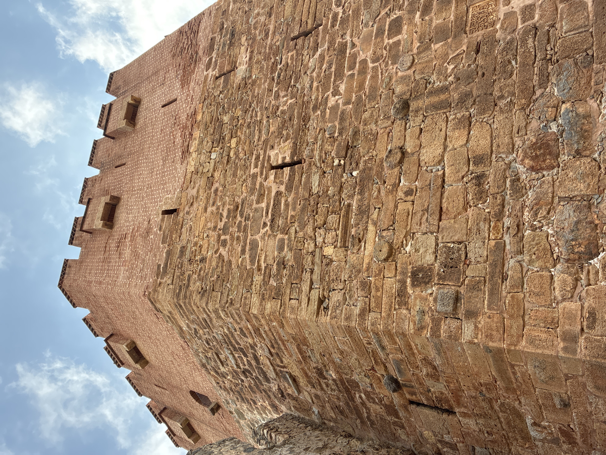 The image shows a low-angle view of the Alanya Red Tower (Kızıl Kule) in Alanya, Turkey. The tower is a historic defensive structure constructed of red brick and stone. The lower portion of the tower features rough-hewn stone blocks, while the upper section is made of finer red brick. The crenellated top of the tower is visible against a sky with scattered clouds. Small, narrow openings, likely arrow slits, are visible on the stone portion of the tower. One of the cropped images reveals a circular window with a metal grate on the upper brick section. Another cropped image shows Arabic inscriptions on a stone section of the tower. The perspective emphasizes the imposing height and robust construction of the tower.