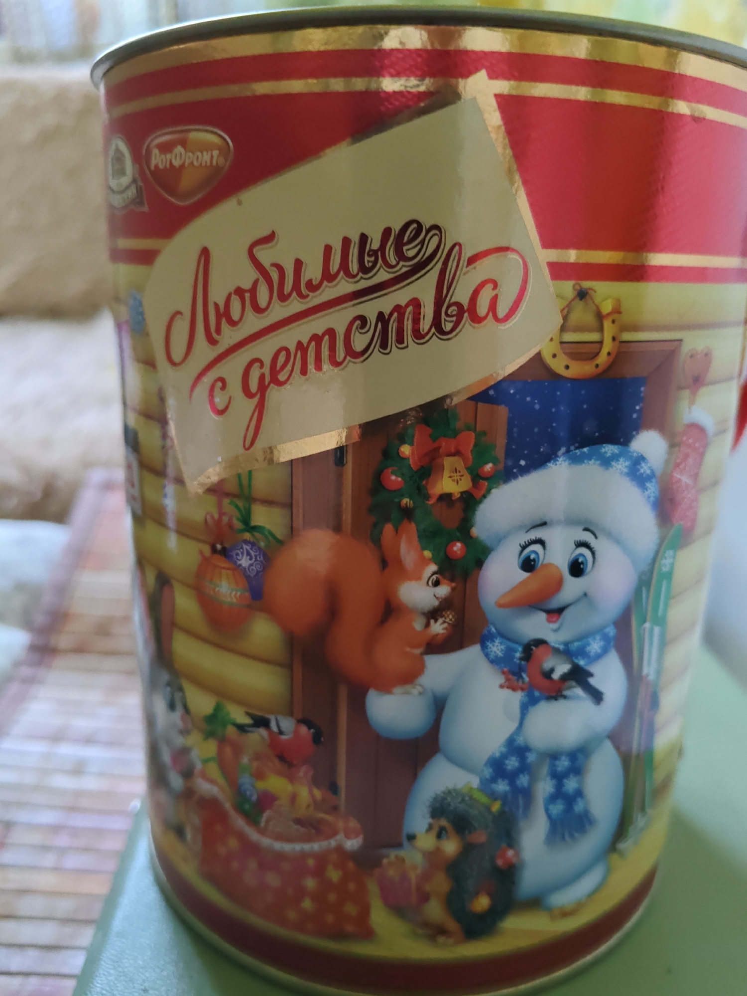 This is a close-up, eye-level photograph of the side of a cylindrical metal tin. The tin features a festive, cartoon-style illustration in a predominantly red and yellow color scheme with gold accents.
The main illustration depicts a winter scene with a smiling snowman wearing a blue patterned scarf and hat. The snowman is holding a small bird with red and black markings on its chest. To the snowman's left is a red squirrel holding a nut. Below the snowman is a hedgehog holding a gift box. Behind the snowman is a doorway leading into a dark interior with a starry sky visible through it. A wreath with a bell hangs on the doorframe. A red Christmas stocking is also visible hanging on the right side of the tin.
A prominent gold sticker with a red, cursive script is placed on the front of the tin, overlaying parts of the illustration. The text on the sticker appears to be in Russian and reads "Любимые с детства," which translates to "Beloved since childhood."
Above the gold sticker, on the red portion of the tin, is a smaller oval logo in gold and red, also with Russian text, which reads "РОТФРОНТ".
The background is out of focus, showing a light-colored, textured surface on the left and a green surface on the right. The overall impression is a decorative tin, likely for candy or cookies, featuring a Christmas or winter theme. There is no indication of the tin being located in Rostov-on-Don, Russia, or any specific city.