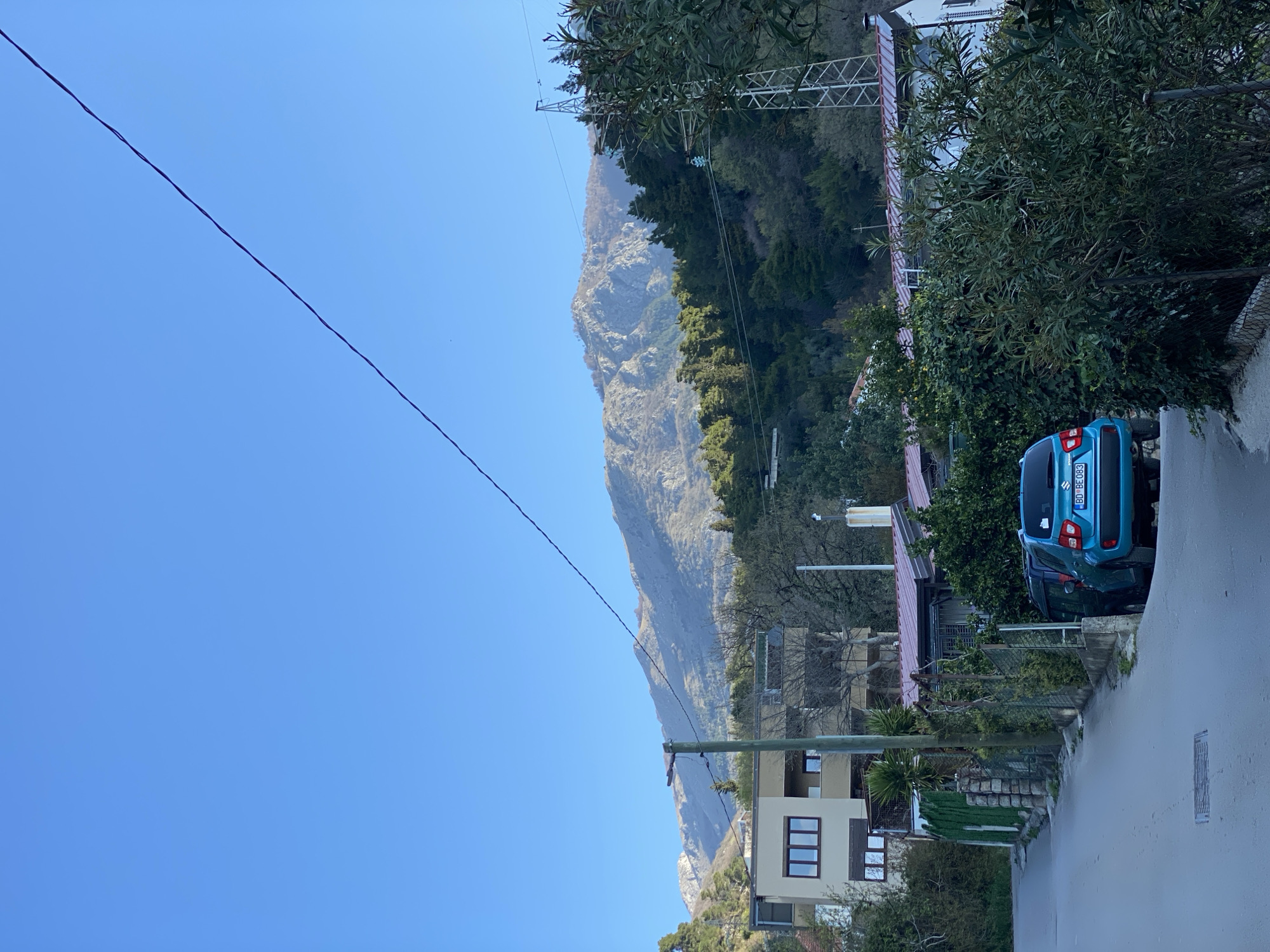 The scene is a street in Budva, Montenegro, showcasing a mountain backdrop, residential structures, and a parked blue car. An overhead power line stretches diagonally across the clear blue sky. The mountain is partially covered in vegetation. A two-story building with windows is visible. A blue car is parked on the side of the road, surrounded by trees and greenery. The road appears wet.