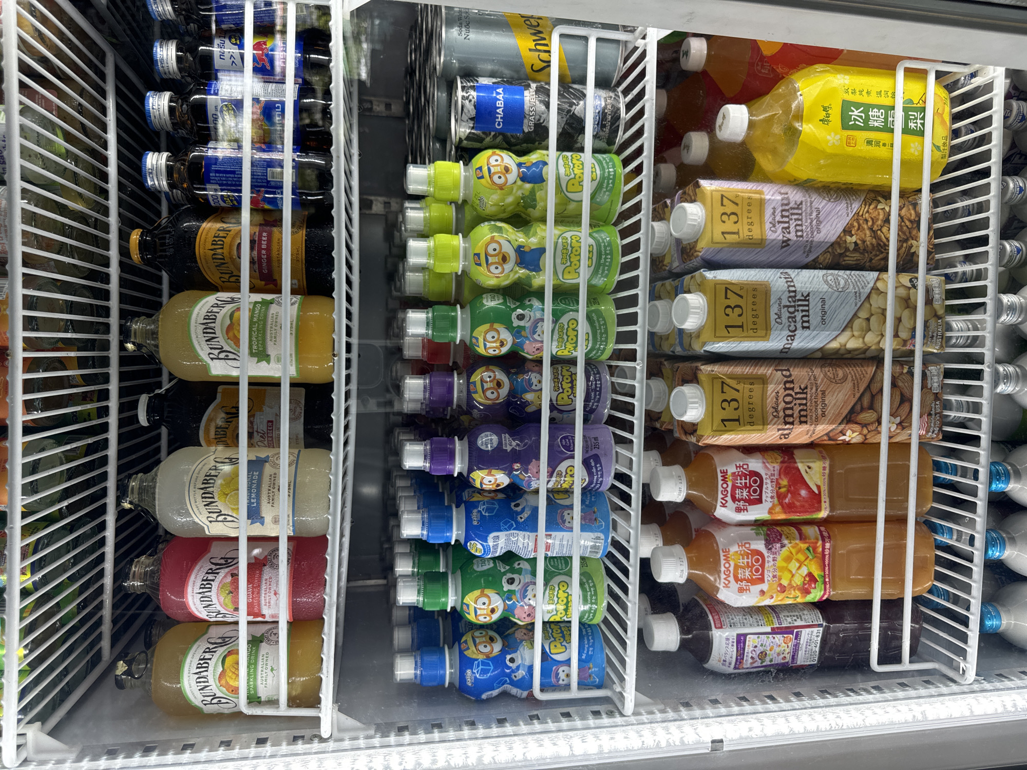 The image depicts a refrigerated display case filled with various beverages. On the left are glass bottles of Bundaberg ginger beer and lemonade in different flavors. The middle section holds plastic bottles of Pororo juice drinks in assorted colors and flavors, and cans of Chabaa grass jelly drink. On the right, there are bottles and cartons of 137 Degrees nut milks, Kagome vegetable juices, and Kang Shi Fu ice sugar pear juice. The bottles are tightly packed on white wire shelves. The location in the city of Đồng Nai, Vietnam is not discernible from the image.