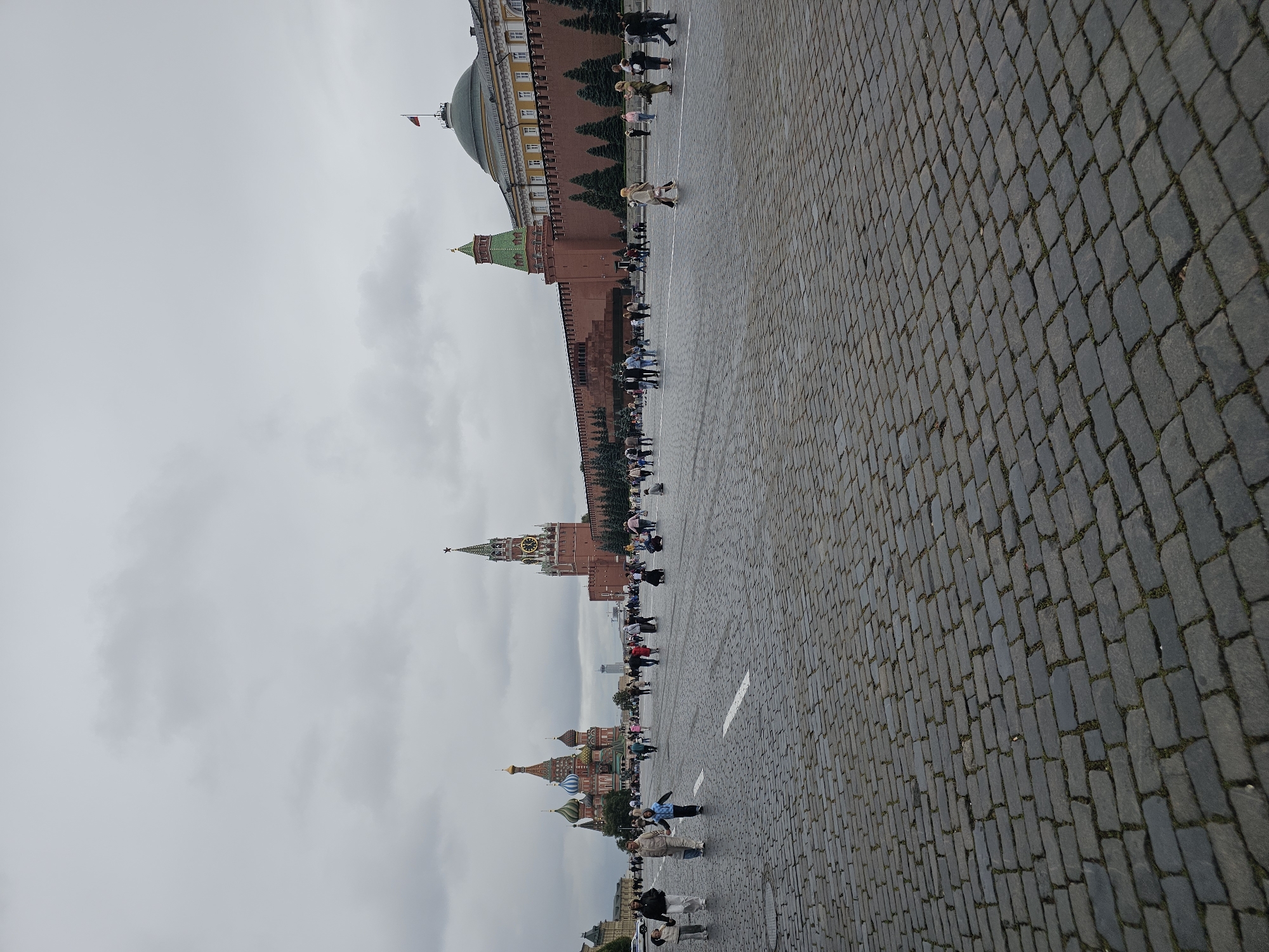 This is a full, wide-angle shot of Red Square in Moscow, Russia, on an overcast day. The foreground is dominated by the cobblestone pavement of the square, with white lines marking pedestrian pathways. In the mid-ground, a crowd of people walks and stands. To the left, the colorful domes of Saint Basil's Cathedral are visible. To the right, the Spasskaya Tower of the Moscow Kremlin, with its clock face, stands tall, and beyond it, the long, red brick wall of the Kremlin, topped by a green-roofed tower and a dome with a Russian flag. The sky is filled with gray clouds.