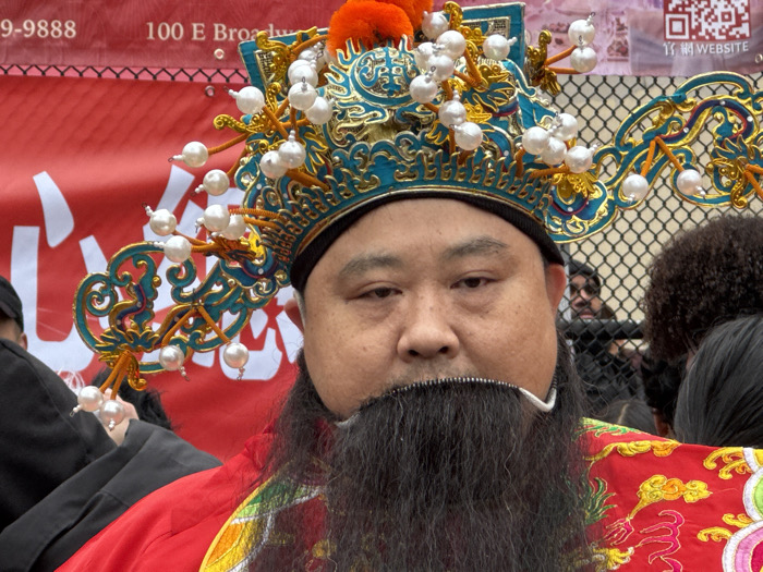 The image features a close-up portrait of a man, likely of East Asian descent, dressed in elaborate traditional attire, positioned centrally in the frame. He has a full, dark black beard that extends down to his chest, with a distinct white or silver U-shaped piece near his mouth, possibly part of the costume's beard. His expression is neutral and direct, looking towards the viewer with slightly narrowed eyes.
He is wearing a striking, ornate headpiece that is predominantly teal or blue-green with intricate gold patterns, possibly depicting traditional motifs like dragons or clouds. The headpiece is adorned with numerous spherical white ornaments, resembling pearls, which are attached by thin, gold-colored wires or stalks that extend outwards from the main structure. On top of the headpiece, there is a small, bright orange pom-pom or tassel. The man is also wearing a vibrant red garment, with intricate gold and multicolored embroidery visible on the shoulder and chest area, featuring floral or symbolic designs.
In the background, a large red banner or fabric is visible, running horizontally across the upper portion of the image. Visible text on the banner includes part of a phone number "9-9888" on the left, "100 E Bro..." (likely "100 E Broadway") in the center-top, and a QR code with Chinese characters (possibly "竹") and the word "WEBSITE" in English on the right.
Behind the red banner, a metal chain-link fence is visible. Through the fence, glimpses of other people can be seen, though they are out of focus. On the right side, a person with dark, curly hair is partially visible, and further back, another person's face can be faintly discerned through the mesh of the fence.
The elaborate costume, particularly the headpiece and beard, suggests the man is participating in a cultural event, parade, or performance, likely related to Chinese traditions. The visible text on the banner, including the address "100 E Broadway," points to a specific location, possibly a Chinese community event in a city like New York, given "E Broadway" is a known street in Chinatown, Manhattan. The scene appears to be taking place outdoors during daylight hours.