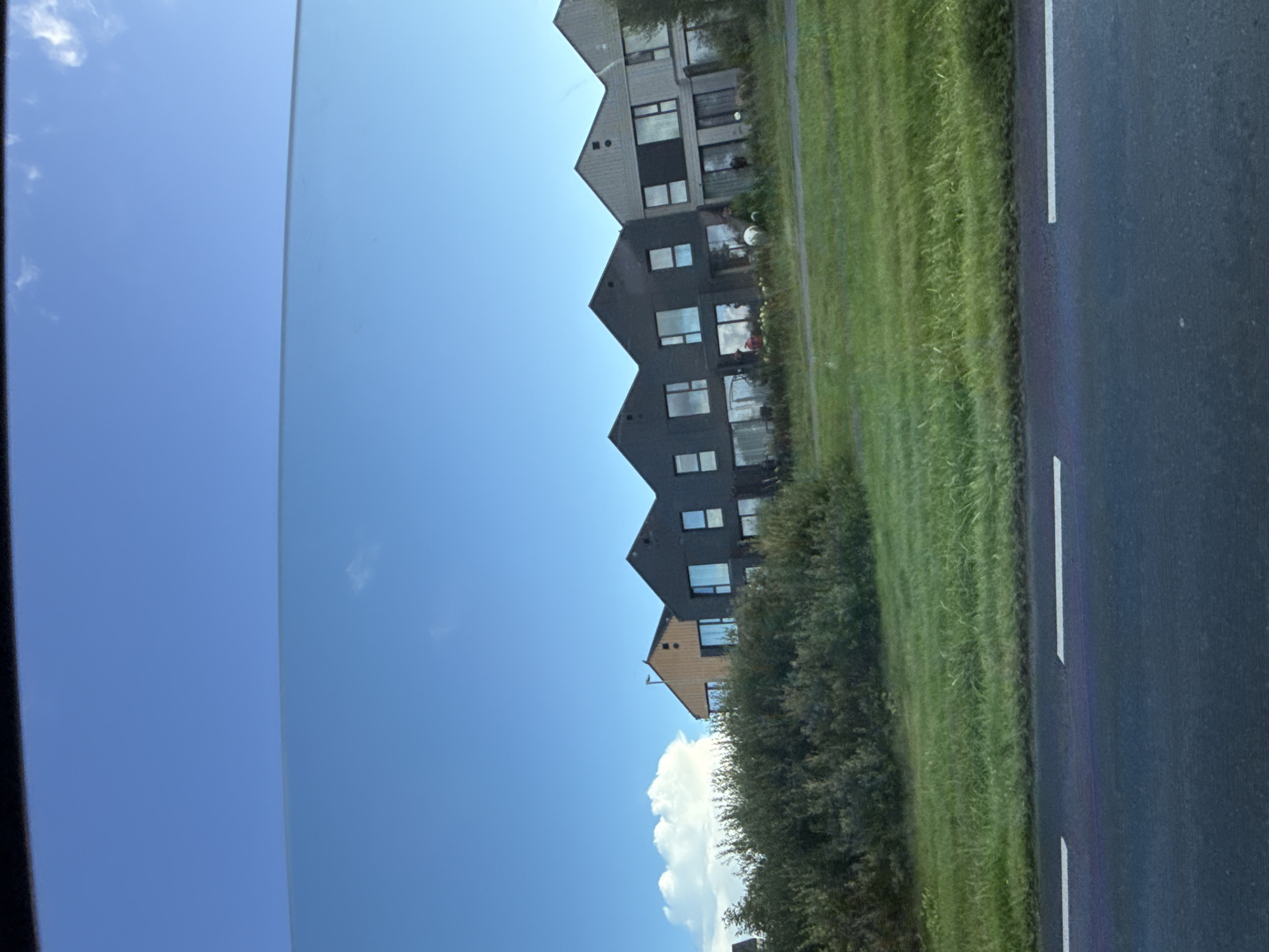 A row of modern, multi-story houses with dark facades and large windows is visible in Harkujärve, Estonia. The houses are situated behind a dense line of green bushes and a grassy slope. The sky above is clear blue with a few small, white clouds. A road with white lane markings is in the foreground.