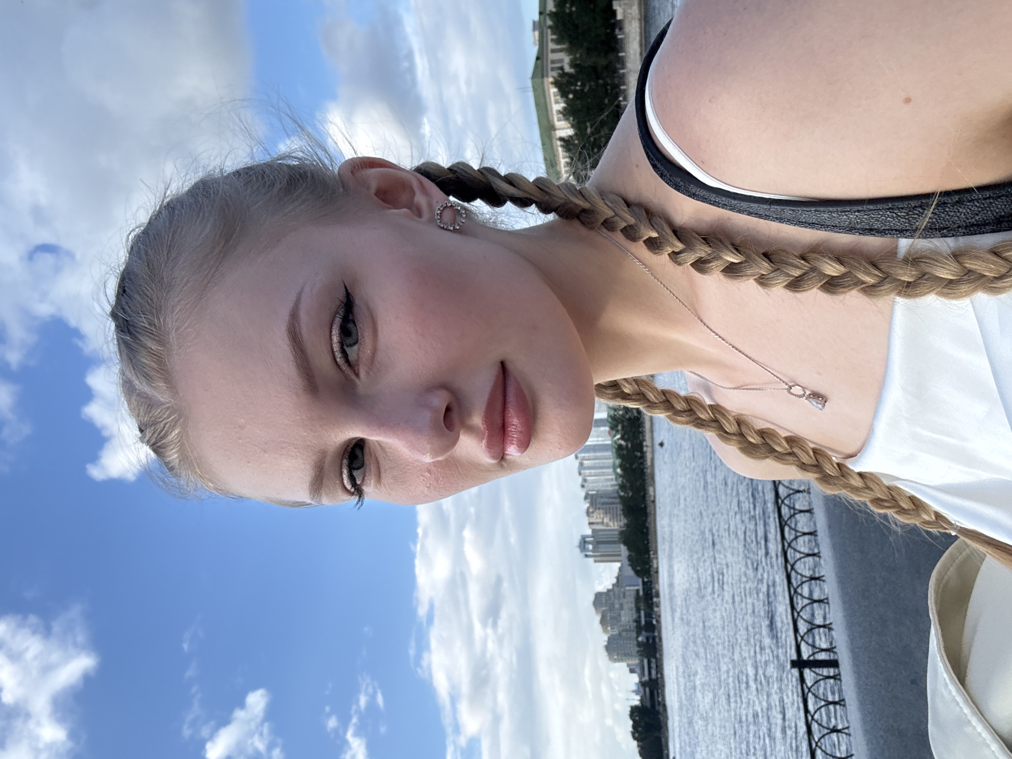 A blonde woman with braided hair is taking a selfie. She is wearing a white top and a silver necklace. Behind her, a body of water is visible, with a city skyline and buildings on the opposite bank. The sky is blue with scattered clouds. The woman is positioned in the foreground, and the background extends to the horizon. The scene appears to be a waterfront area in Yekaterinburg, Russia, likely along the Iset River, given the urban landscape and the presence of a body of water.