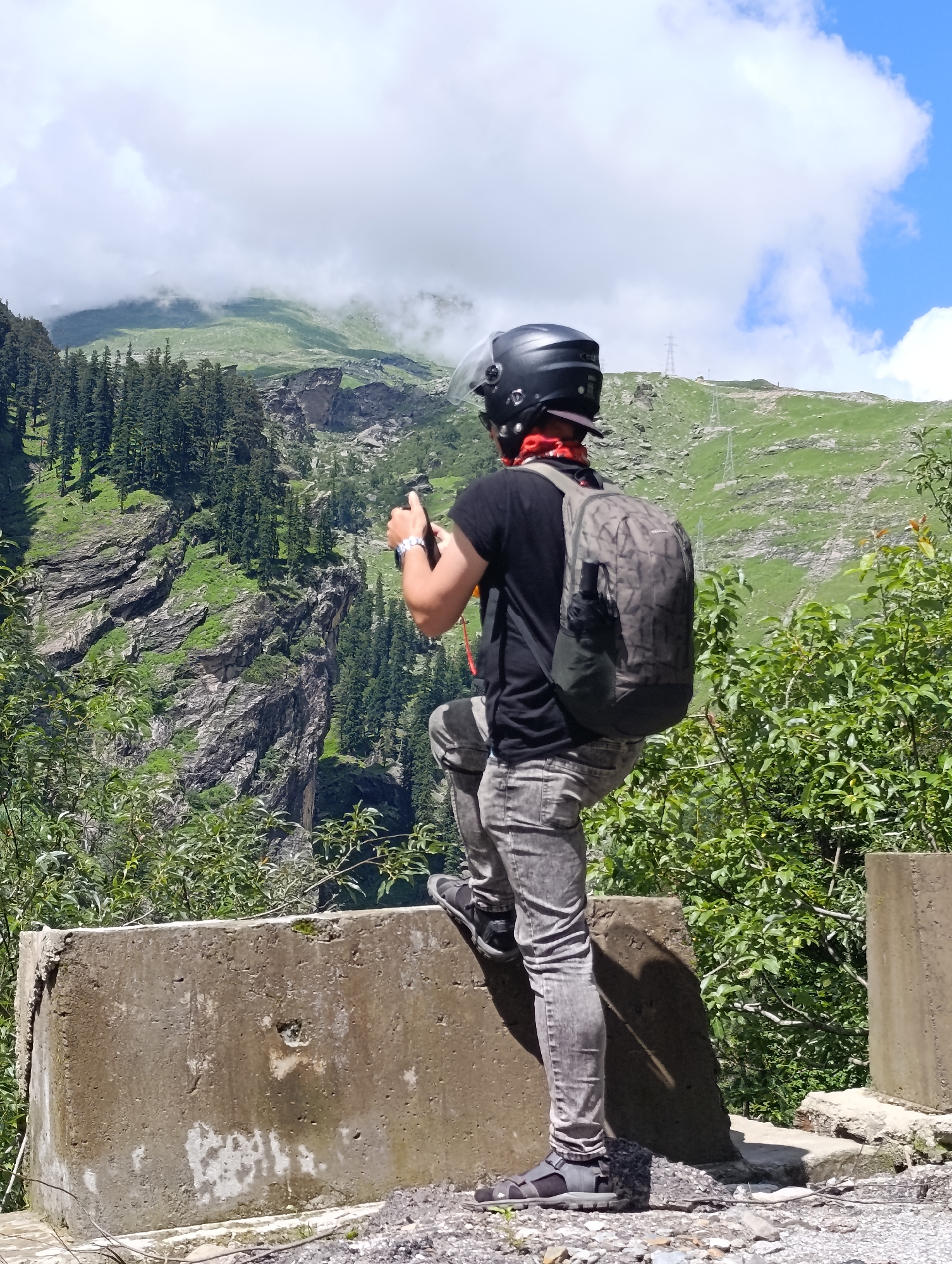 In an outdoor, mountainous scene, a person wearing a black t-shirt, grey pants, a black helmet, and a patterned backpack stands with their right foot on a concrete barrier, looking towards a steep, rocky mountain slope covered in green vegetation and trees. The person is holding a mobile phone in their right hand. The sky is blue with scattered white clouds.