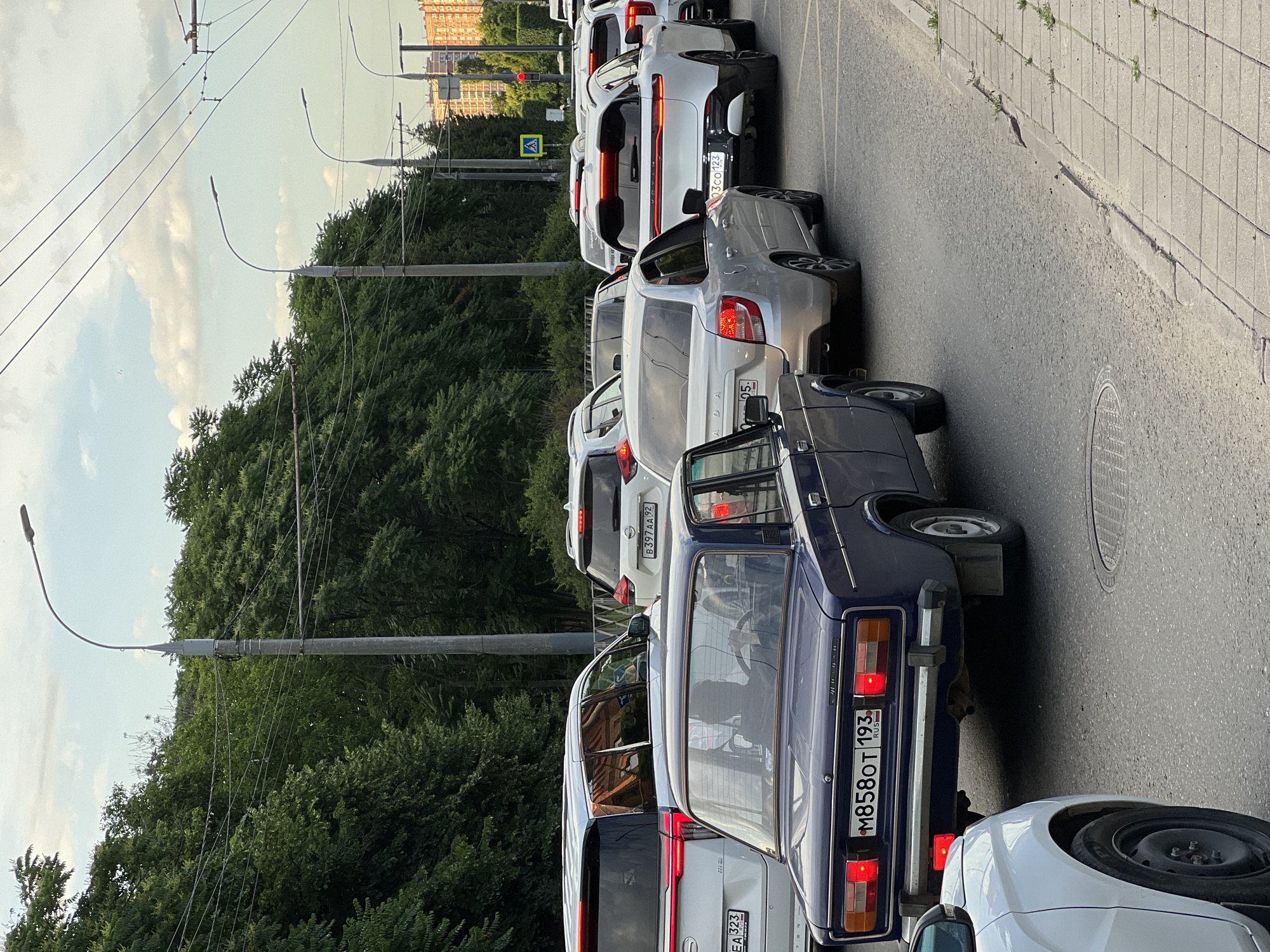 The image depicts a traffic jam on a city street, likely in Krasnodar, Russia, given the license plates. A line of cars is stopped on the asphalt road, with a pedestrian sidewalk visible on the right. The cars are of various models and colors, including a dark blue sedan in the foreground with a license plate reading "M858OT 193 RUS". Behind it, a silver sedan and several white SUVs are visible. Trees line the left side of the road, and power lines are strung overhead. In the background, buildings and more trees can be seen. The sky is partly cloudy. A traffic light is visible in the distance, appearing red, indicating a stop. A pedestrian crossing sign is also present. No individuals are clearly identifiable in the vehicles, except for a silhouette of a person behind the steering wheel of the blue sedan. The overall scene suggests a typical urban traffic situation during daylight.