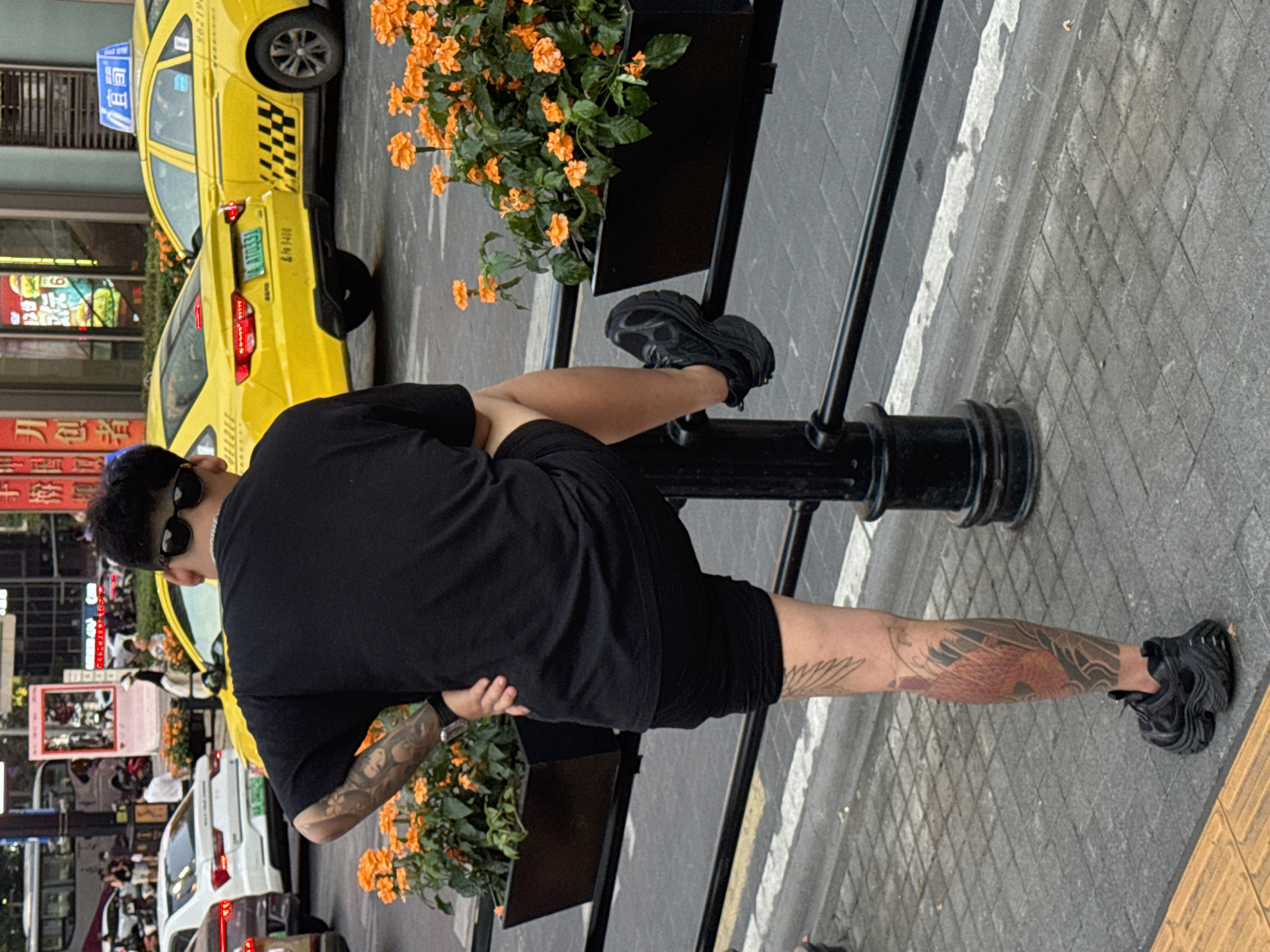 A person with multiple tattoos is standing on a sidewalk in the Yuzhong District of China, with their right leg resting on a metal railing. The person is wearing a black t-shirt, black shorts, and black sneakers. They have sunglasses on and a watch on their left wrist. In the background, there are yellow taxis, cars, pedestrians, and buildings. There are also planters with orange flowers. The scene appears to be a street in a busy urban area.