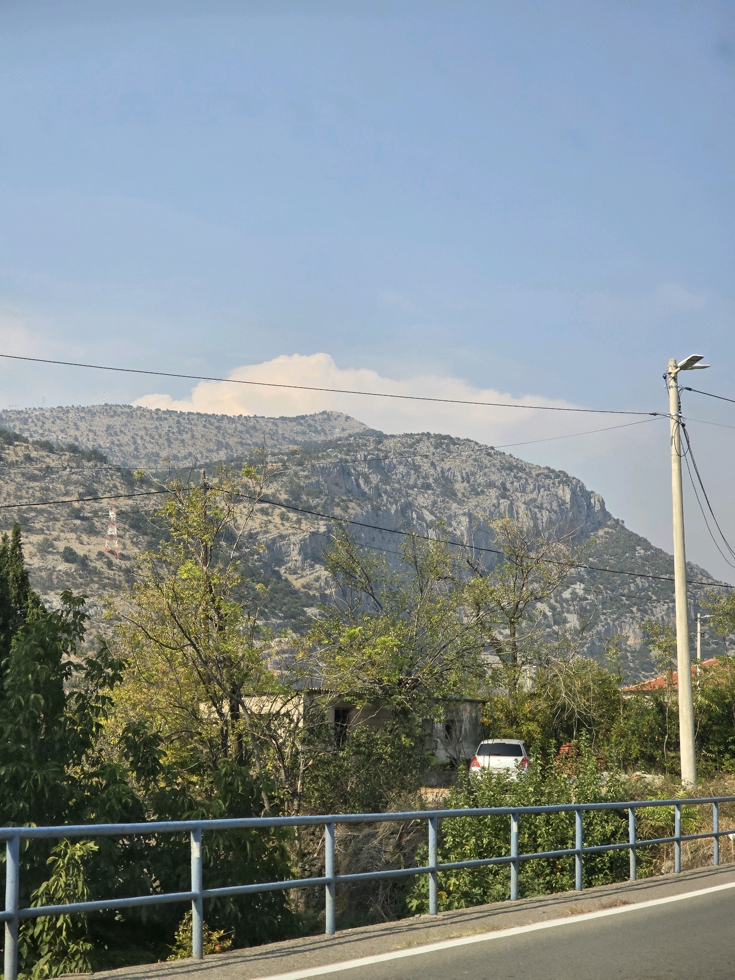 This is a landscape image taken in Podgorica, Montenegro. In the foreground, a grey asphalt road with a white line is visible, bordered by a blue metal railing. Beyond the railing, lush green trees and bushes dominate the middle ground. A white SUV is parked among the foliage, partially obscured by branches. Behind the vegetation, an old, weathered building stands. In the background, a large, rocky mountain range rises under a clear blue sky with scattered white clouds. A red and white communications tower is situated on the side of the mountain. A concrete lamppost with a modern lamp fixture stands to the right of the frame. Overhead, dark power lines stretch across the sky.