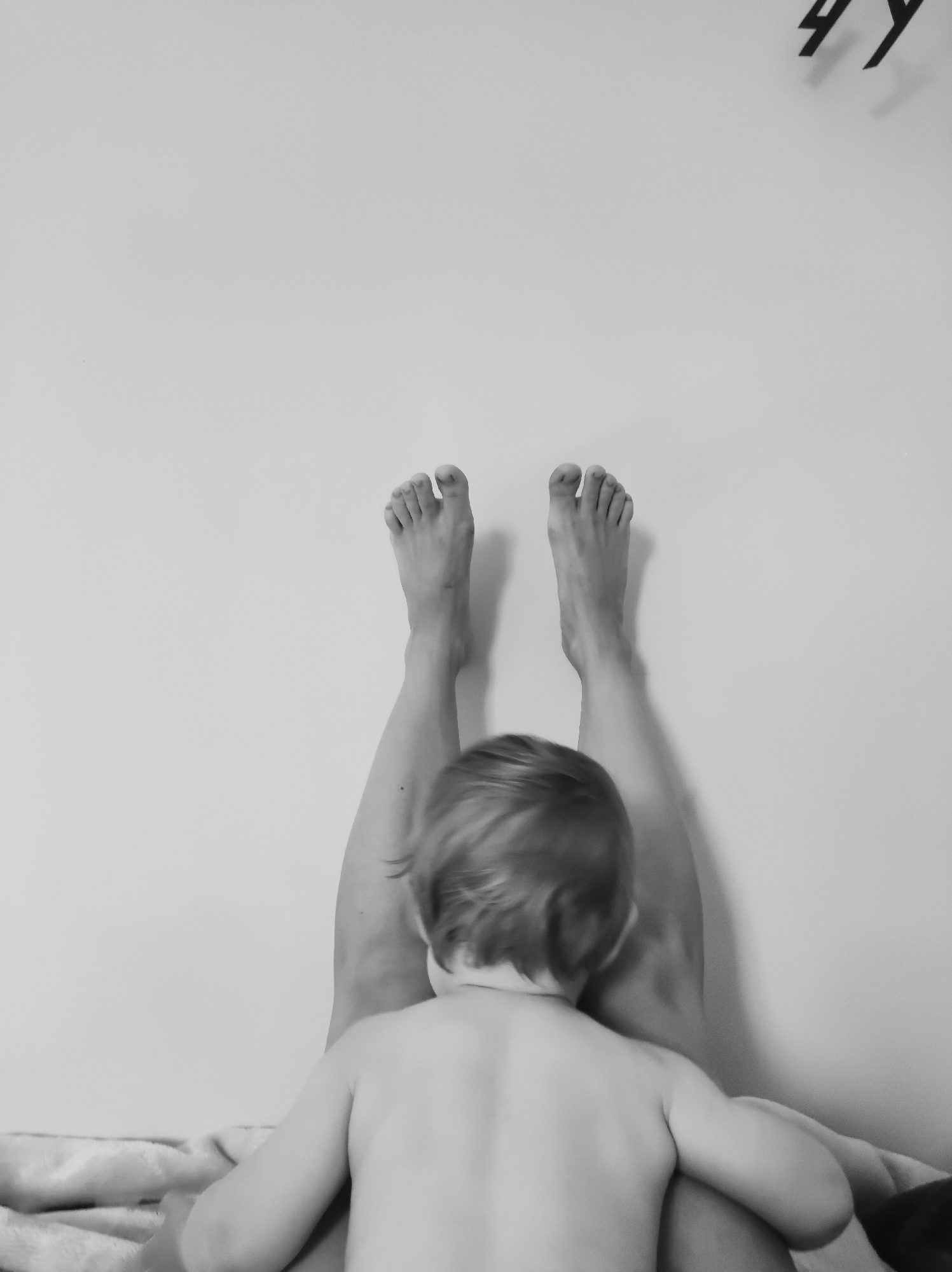 A black and white, eye-level shot shows a child lying against a woman's legs, which are extended up against a wall. The child, who is viewed from behind, has short hair and is shirtless. The woman's legs are straight and extend upwards, with her feet near the top of the frame. The background is a plain wall, with a dark, abstract object hanging on it in the upper right corner. A blanket is visible under the child and woman's legs.
The image does not appear to be set in the city of Фоки, Russia. The scene is indoors, likely in a home.