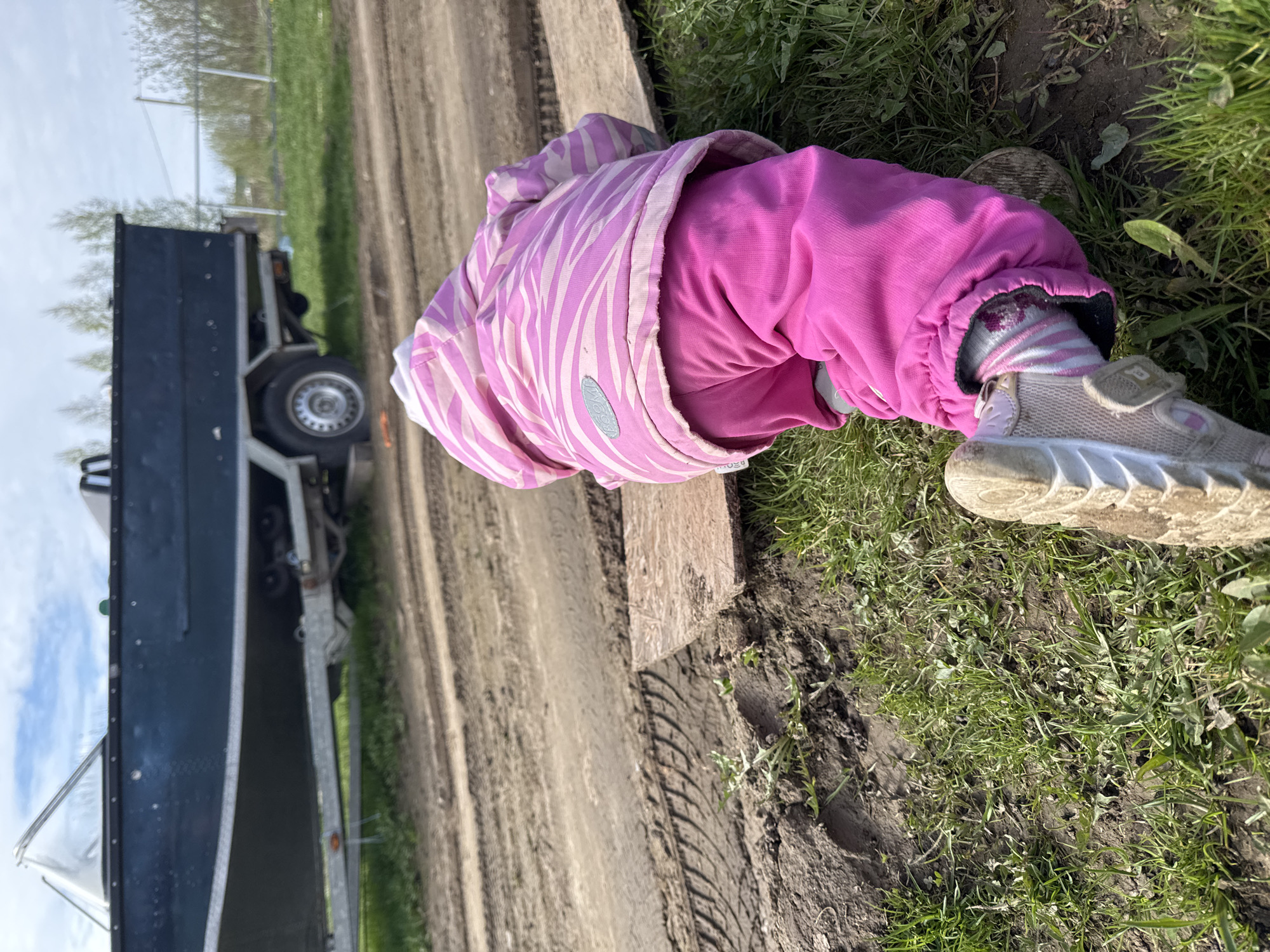 A child lies prone on the ground in Тоншалово, Russia, near a dirt road and a parked trailer. The child wears a pink and white striped jacket, pink pants, striped socks, and light-colored shoes with a velcro strap. The ground is a mix of dirt and green grass, and a piece of plywood lies between the child and the dirt road. The trailer, attached to a vehicle, is visible in the background on the left side of the image. The weather appears to be clear and sunny.