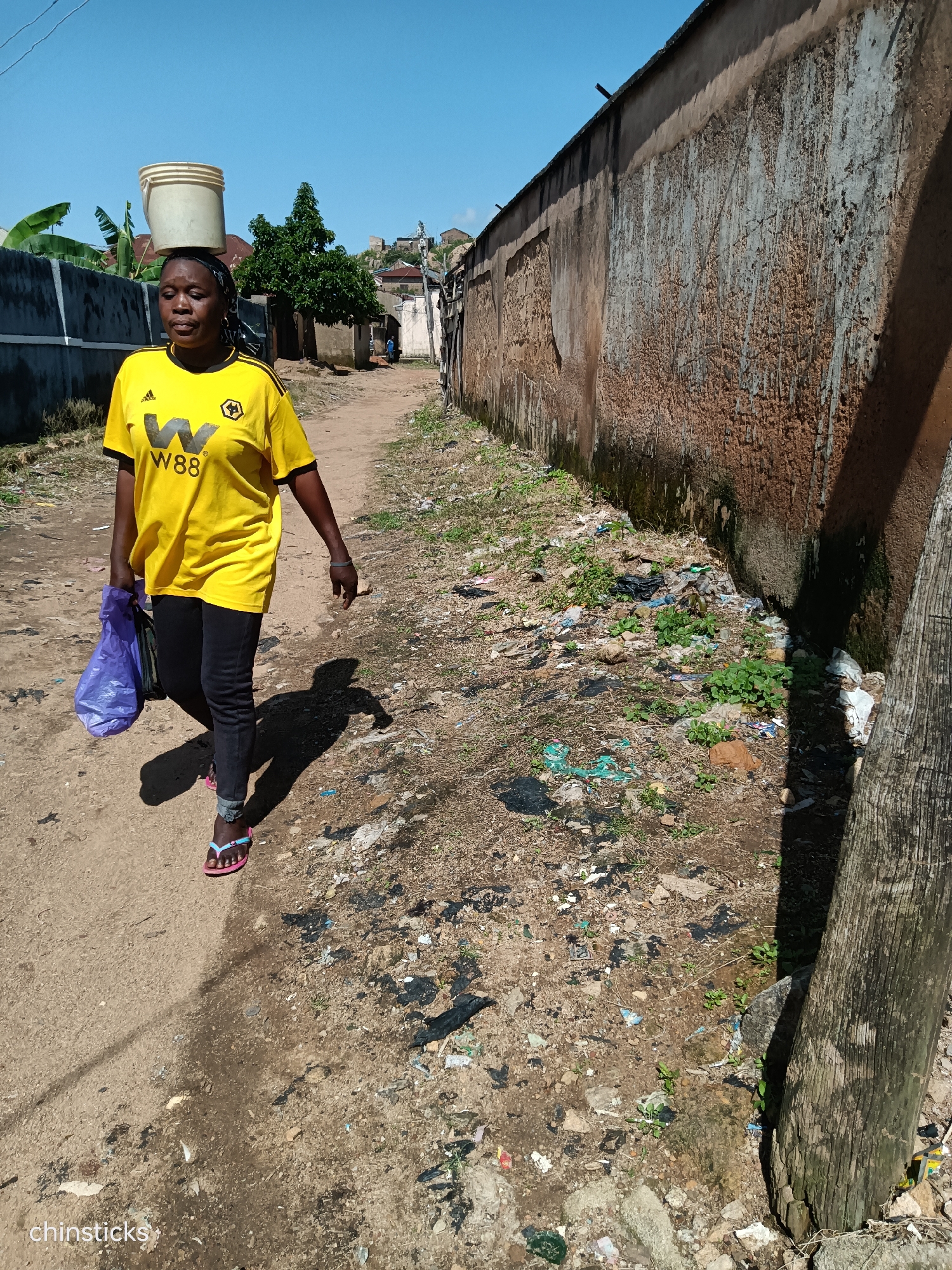 A woman walks down a dirt path in Jos, Nigeria, balancing a bucket of water on her head and carrying a purple plastic bag in her left hand. She is wearing a bright yellow t-shirt with a "W88" logo and black leggings, along with pink flip-flops. The path is lined on the right by a rough, textured, ochre-colored wall. The ground is littered with various debris, including plastic bags, food wrappers, and plant matter. Green vegetation is growing sporadically among the trash. On the left side of the path, a lower concrete wall is visible, with more greenery behind it. The sky above is a clear blue. The overall impression is a scene of everyday life in a somewhat unkempt urban environment.