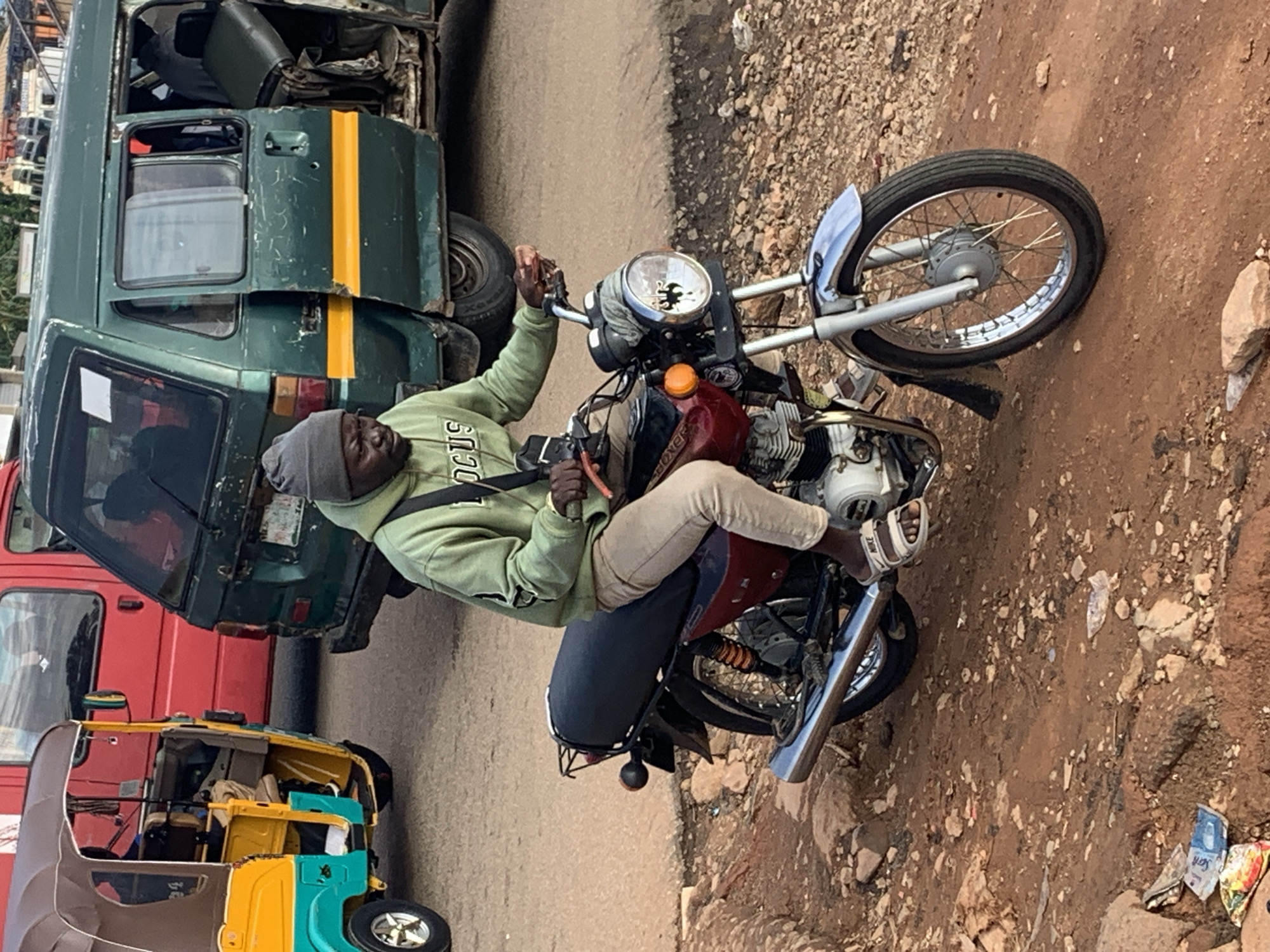 A man wearing a grey beanie and a green hoodie with "FOCUS" printed on it, is riding a red and black motorcycle down a dirt road. He is wearing beige pants and white sandals. The motorcycle has a chrome exhaust pipe and a circular headlight. In the background, a green van with open doors and a red bus are parked. A yellow and blue auto-rickshaw is visible to the left. The road is covered in dirt and small stones. The scene appears to be in Gura Top, Nigeria.