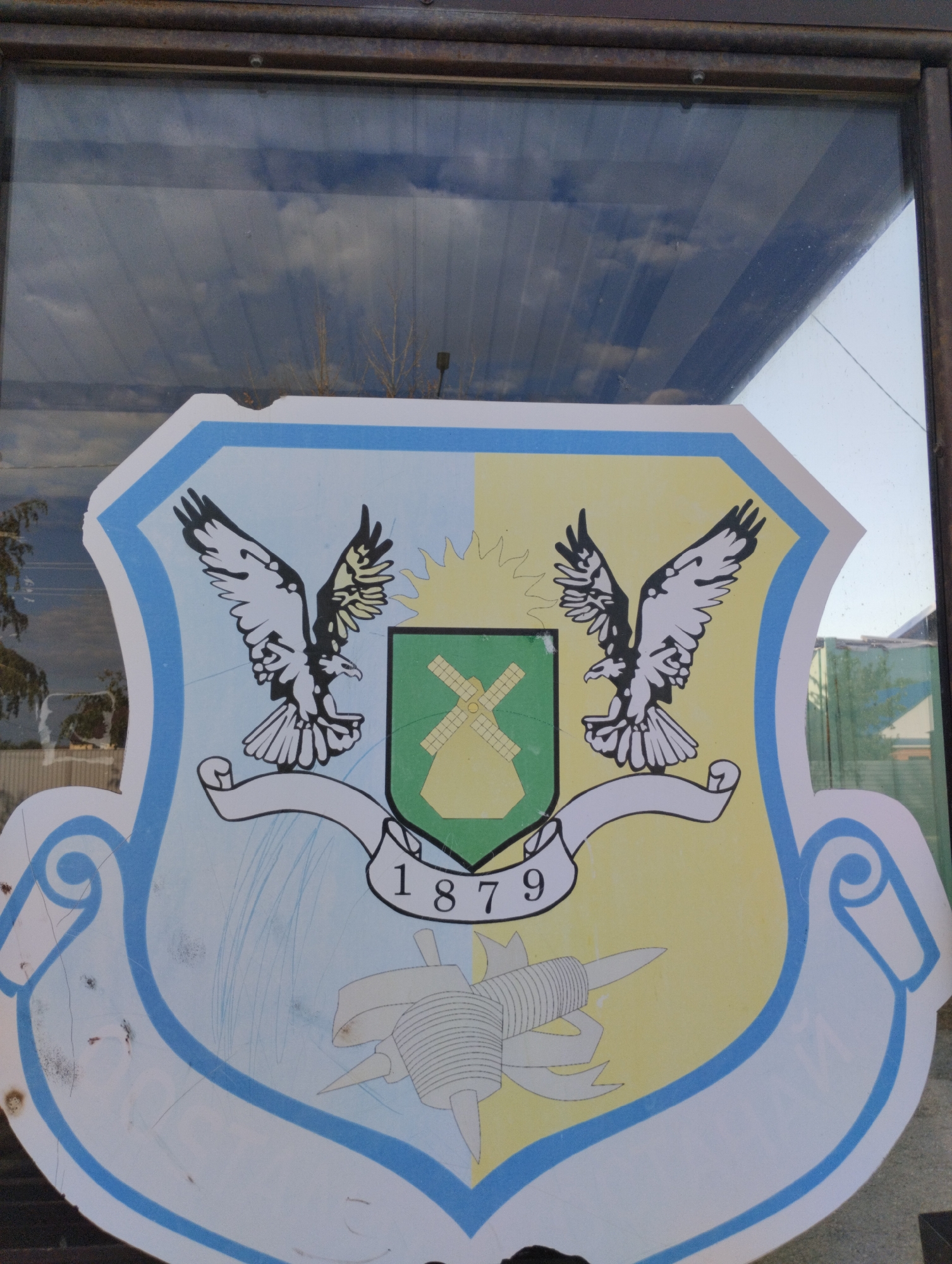 The image shows a crest with two eagles on either side of a shield. The shield is divided into a light blue and yellow halves. On the light blue half, there is a drawing of a windmill with what appears to be a sunburst behind it. On the yellow half, there are no distinct symbols. Below the shield is a banner with the numbers "1879" written on it. The crest has a light blue border. There is a drawing of a stylized object below the banner. The crest is displayed on what appears to be a glass surface, with reflections of the sky and trees visible. This crest is located on the building of the Regional Assembly of People of Kazakhstan in Kostanay, Kazakhstan.