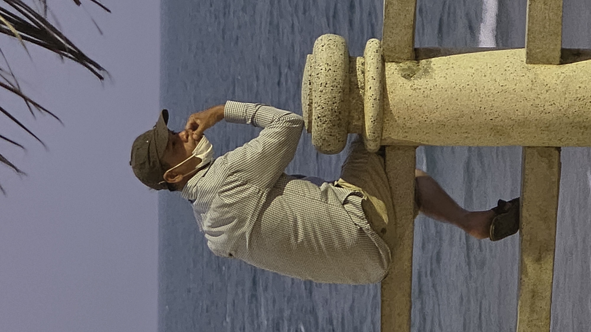 A man is sitting on a stone structure in Nha Trang, Vietnam. He is wearing a baseball cap, a checkered long-sleeved shirt, khaki shorts, and a face mask. He is holding his hand to his face. The stone structure is a part of a decorative fence, with the sea visible in the background. Palm fronds are visible in the upper left corner.