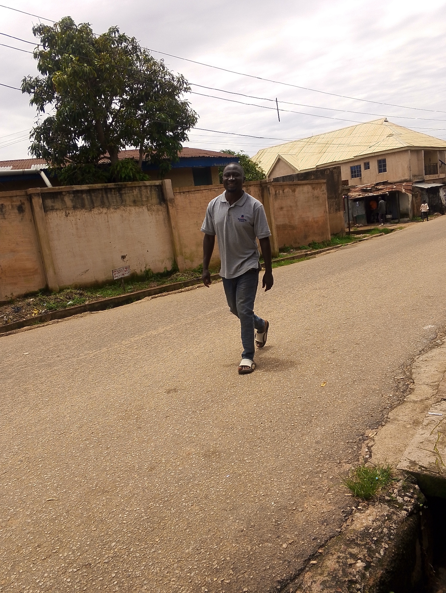 A medium shot captures a man walking on an asphalt road in a cloudy outdoor environment. The man is wearing a grey polo shirt with a logo and blue jeans, and he has on white slide sandals. He is walking towards the viewer with a slight smile. Behind him, to the left, is a light-colored concrete wall with some graffiti and a tree with green foliage. To the right of the man and the wall, there are several buildings, one with a light yellow roof and the other appears to be a smaller structure with people gathered. The road has some cracks and a drainage ditch is visible on the right side of the image. The sky is overcast, suggesting a dull day.