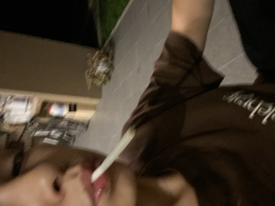 A close-up, blurred, angled image shows a person with a pale tongue sticking out, holding a white object (possibly a cigarette) between their lips. They are wearing a brown shirt. In the background are elements of a brick building, sparse vegetation, and what appears to be a cluttered outdoor space with boxes or materials, all under dim lighting. The image is low quality and lacks sharpness.