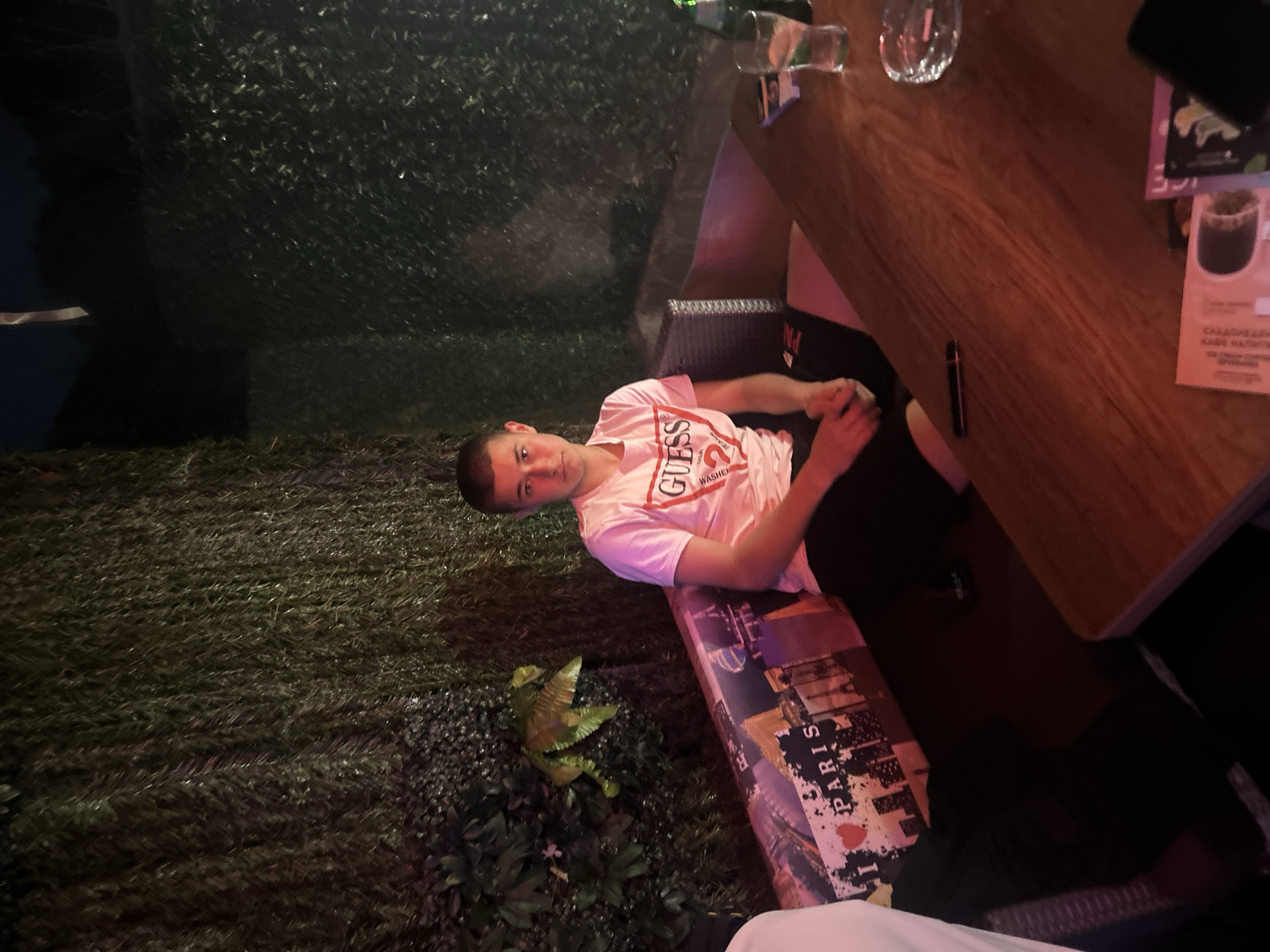 A young man sits at a table in an outdoor setting. He wears a white Guess t-shirt and dark pants. The table is wooden and has glasses, menus, and a black object on it. The background includes artificial greenery and a bench with a Paris-themed design. The location is possibly in Elhovo, Bulgaria.