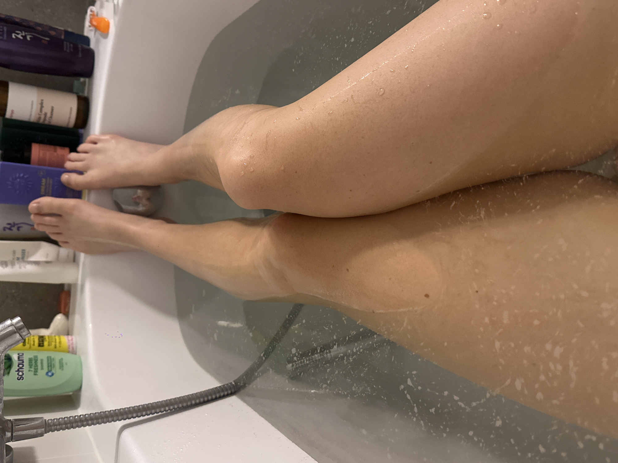 The image shows a person's legs submerged in a bathtub filled with water. The water has some foam and bubbles. The person's legs are the primary focus, with water droplets visible on their skin. Various toiletries and bathing products are lined up on the edge of the bathtub in the background. There is no indication of the location in Krakow, Poland.
