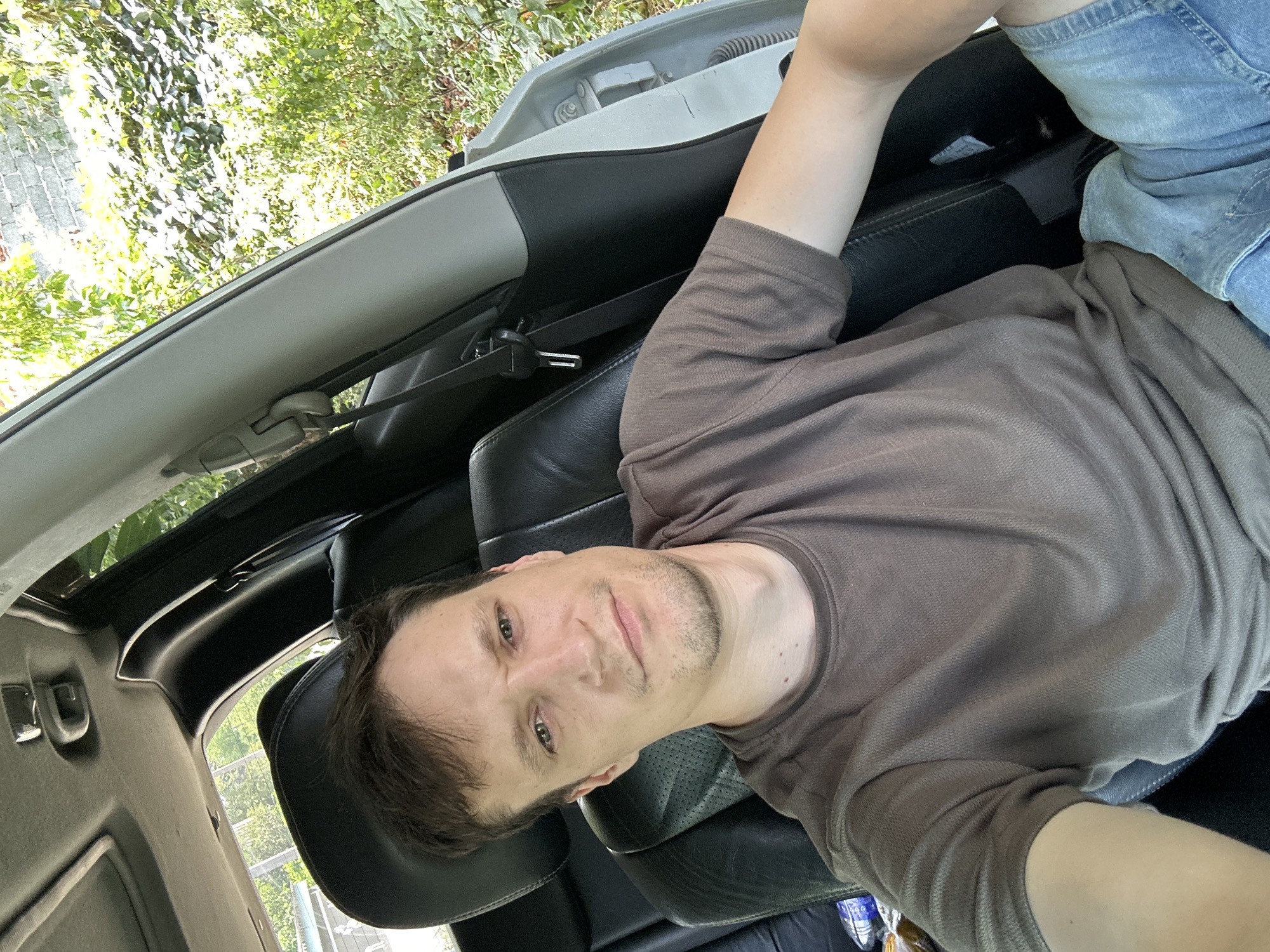 A man is sitting in the driver's seat of a car, looking at the camera with a neutral expression. He is wearing a brown t-shirt and blue jean shorts. The car appears to be parked, with the door open. Outside the car, there is lush green foliage and a brick wall visible. The interior of the car is black leather. The scene is likely outdoors, possibly in a residential area or a park, given the vegetation. The image is a selfie taken from a slightly high angle.