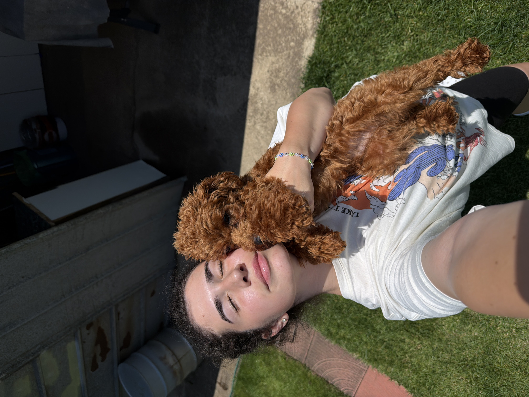 This is a high-angle, full shot of a young person lying on the grass, with a small, fluffy, reddish-brown dog resting on their chest. The person is looking upwards with their eyes closed and a slight smile, appearing relaxed and content. The dog is nuzzling into the person's face, also looking peaceful. The person is wearing a white t-shirt with an orange and blue graphic design and black shorts. A colorful beaded bracelet with green, blue, and white beads is visible on the person's left wrist.
The scene is outdoors on a sunny day, with bright sunlight casting shadows on the concrete ground to the left of the frame. The grass is green and well-maintained. In the background, there are some weathered wooden structures and what appears to be industrial or storage elements, suggesting a backyard or an area with some utilitarian features. The lighting indicates it is likely midday. The overall mood is one of warmth, affection, and tranquility, highlighting the bond between the person and the dog.