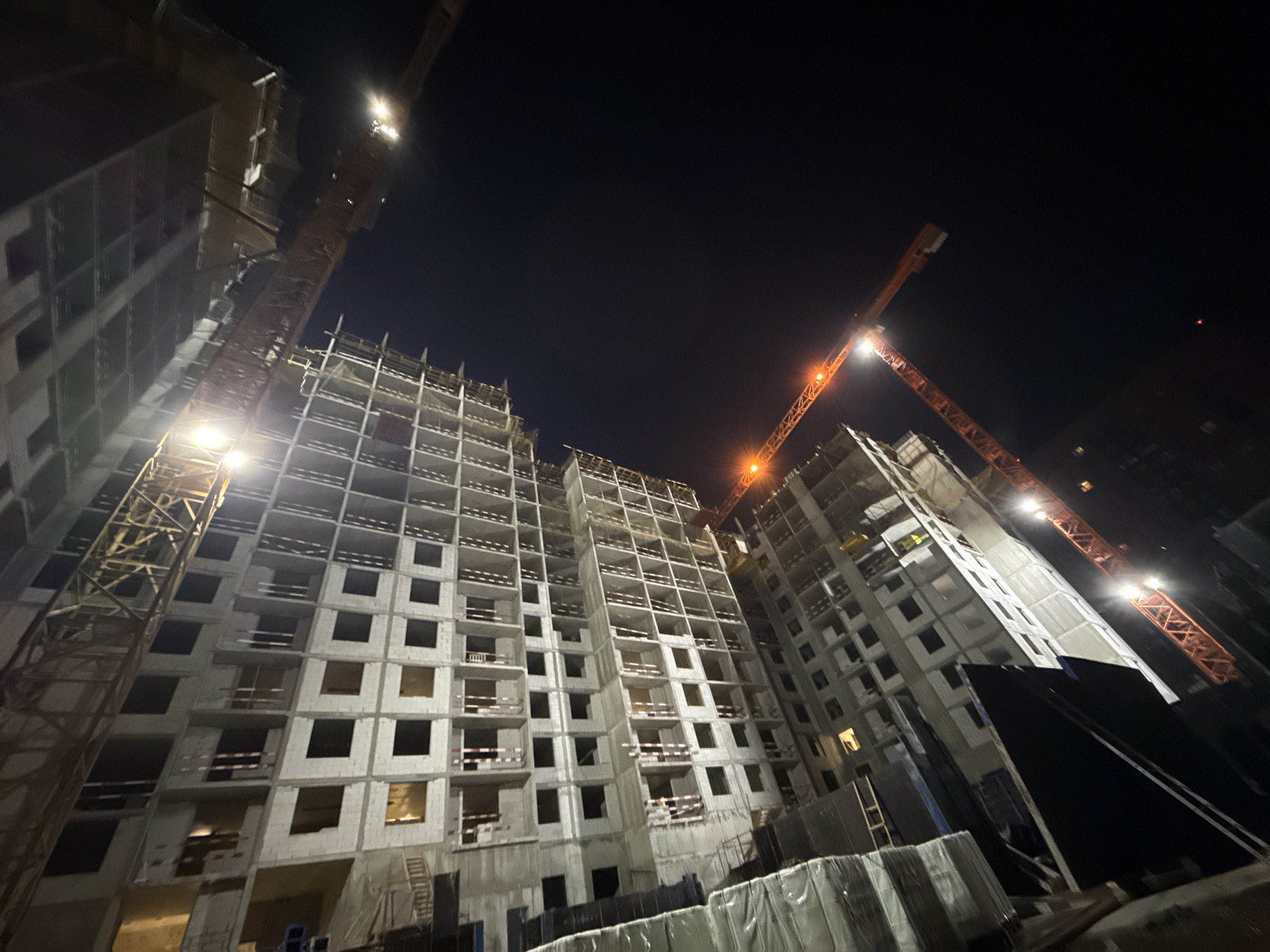 This is a low-angle, night-time photograph of a large-scale construction site in Moscow, Russia. Two massive orange tower cranes are prominently visible, their illuminated booms extending over partially constructed high-rise buildings. The buildings themselves appear to be residential apartment blocks, constructed from concrete and rebar, with numerous unfinished window openings. The scene is stark and industrial, with lights from the cranes and some internal building lights illuminating the structures against the dark night sky. Construction materials, possibly wrapped in plastic sheeting, are visible in the foreground. The overall impression is one of ongoing development in an urban environment.