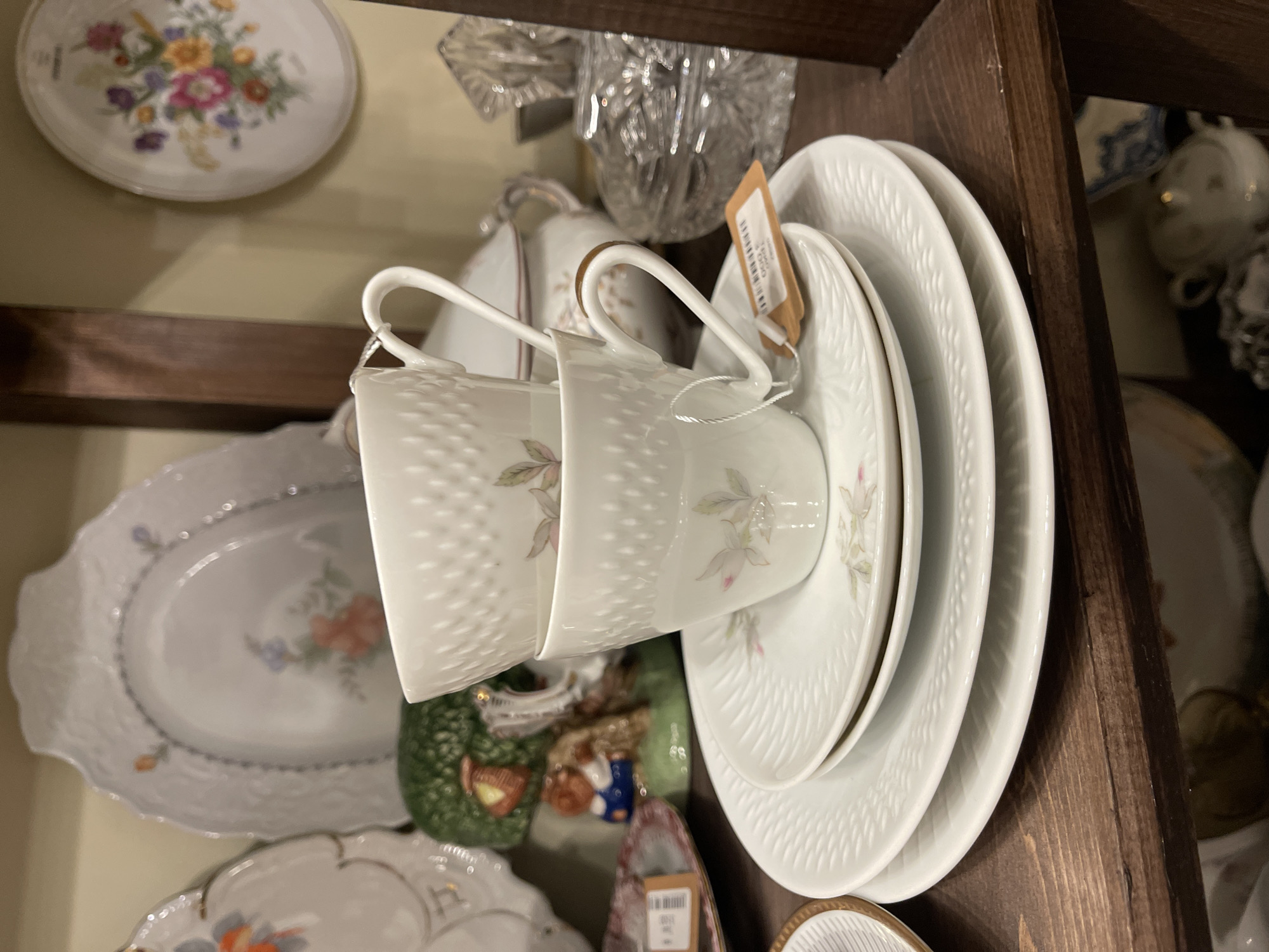 The image depicts a close-up view of a shelf in what appears to be a shop or a home display, showcasing antique or decorative porcelain items. In the foreground, a stack of white plates with intricate embossed patterns is visible, topped with two matching teacups, one slightly nested inside the other. The teacups have a similar textured exterior and delicate floral decals on the sides. A small brown tag with text is attached to the handle of the bottom teacup.
In the background, to the left, parts of other porcelain pieces are visible, including a large, ornate serving dish with a floral motif and a small decorative figurine of a person in blue pants near a green, tree-like object. To the right, a cut-glass crystal object and a lidded porcelain container are on the same shelf. Further in the background, another decorative plate with a colorful floral design is displayed on the wall. The shelf itself is made of dark wood, and the overall lighting suggests an indoor setting, possibly a curio shop or an antique store.
The provided context specifies the location as Svetlogorsk, Russia. This detail, however, cannot be visually confirmed from the image content itself. The scene is static, with no people or actions depicted, focusing solely on the arrangement of decorative porcelain objects.