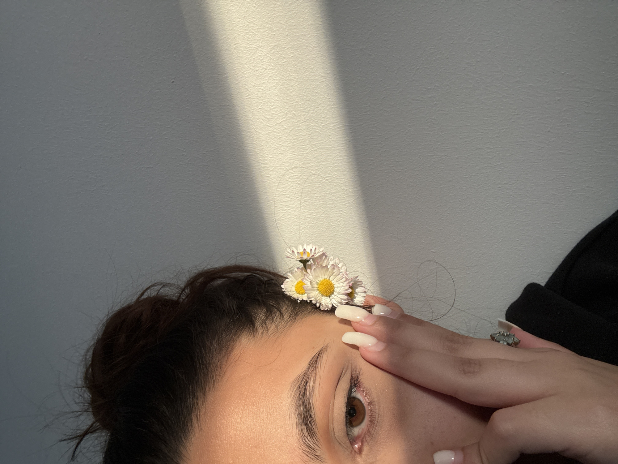 The image shows the upper part of a person's face, angled upwards. Daisies are resting on the forehead. The person's hand is placed on their forehead near their eye. A ring is visible on one of the fingers. The background is a white wall with a streak of sunlight. There is no information in the image to suggest the location is Mönchengladbach, Germany.