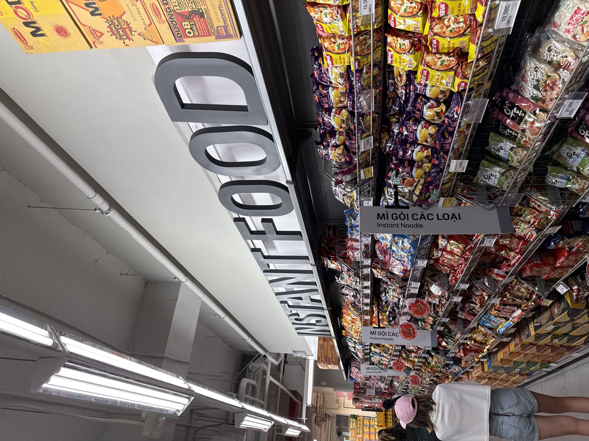 The image depicts the interior of a supermarket aisle in Nha Trang, Vietnam, with a focus on the "Instant Food" section. Large, black, block letters spelling out "INSTANT FOOD" are prominently displayed on a white wall above shelves stocked with various instant noodle products. The shelves are organized with numerous colorful packets of instant noodles, some with visible pricing. In the foreground, a young woman wearing a white crop top, denim shorts, and a pink baseball cap is walking away from the camera, down the aisle. Other shoppers are partially visible in the background. The lighting appears to be from overhead fluorescent fixtures. The overall environment suggests a typical retail setting for groceries, specifically focusing on convenience food items.