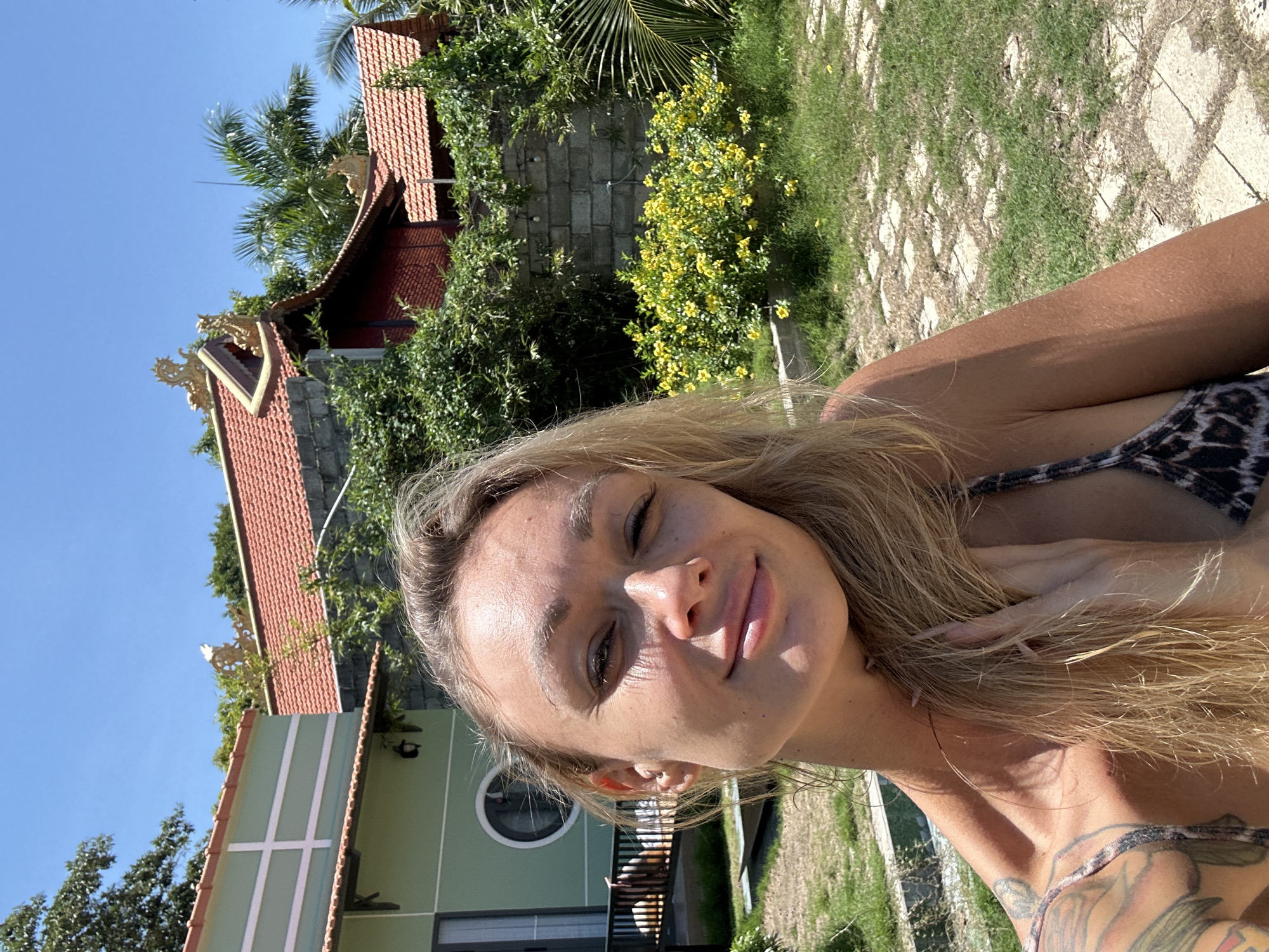 A sideways close-up shot shows a blonde woman with fair skin in Mũi Né, Vietnam, with architectural and natural surroundings. The woman is in the foreground; her fair skin shows some sun exposure, and she has several tattoos on her upper chest and shoulder. She is wearing a bikini top with a leopard print. Her blonde hair is long and slightly tousled.
In the background, there is a building with a multi-tiered roof, red tiles, and decorative golden accents. The architecture is set amidst green foliage, including trees and flowering shrubs with yellow blooms. A stone wall is also visible within the greenery.
To the right of the building, there is a view of a green lawn dotted with stone slabs forming a path.
The sky above is clear and blue, indicating a sunny day.