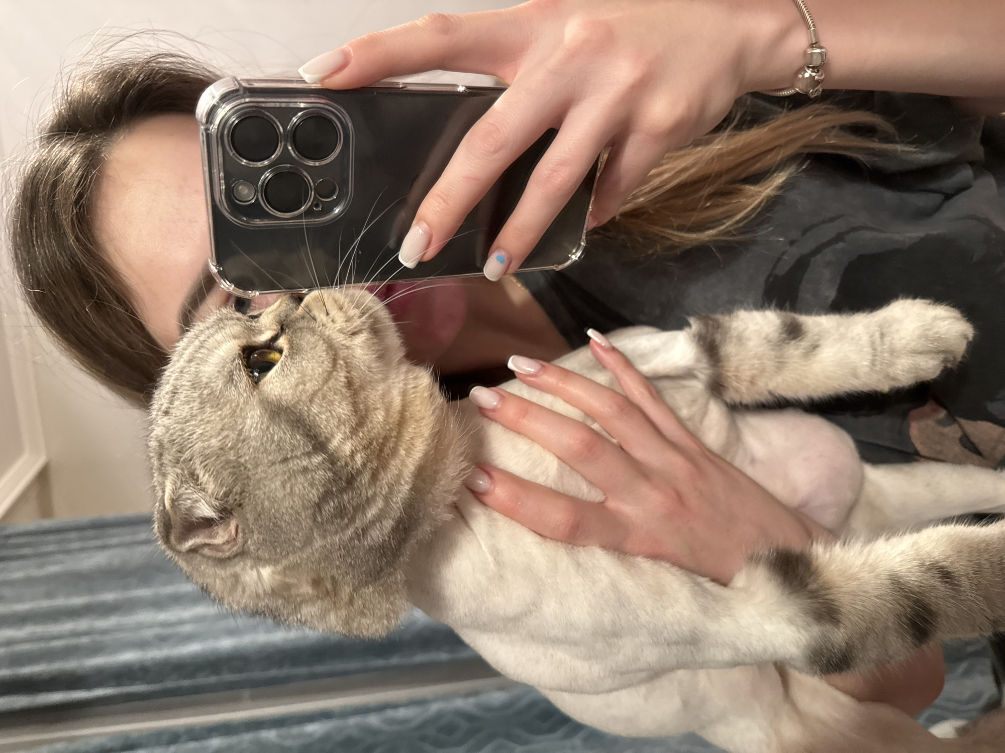 A person is holding a tan and white tabby cat and taking a photo of it with a smartphone. The phone has a clear case with multiple camera lenses. The person's left hand is holding the phone and cat. The cat's face is visible near the phone. The person's right hand is cradling the cat. The person has long white nails with a blue dot on one nail. The person is wearing a silver bracelet. The cat has very small, folded ears. The cat is looking up at the phone. The background is blurry and appears to be indoors.