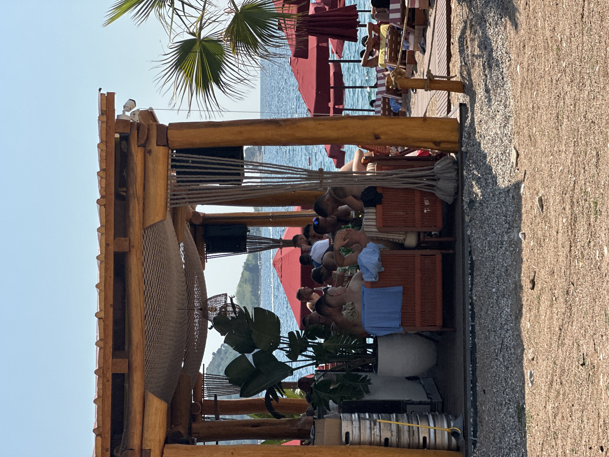 The image depicts a bustling beach bar in Budva, Montenegro, under a clear blue sky. The foreground shows a gravel and sandy beach area with scattered small stones. In the middle ground, a wooden, open-air structure with a thatched roof provides shade for patrons. Inside this structure, several people are seated at tables, some appearing to be engaged in conversation and enjoying drinks. Large potted plants add a tropical feel to the setting. To the right of the structure, a pathway made of wooden planks leads towards the beach.
Beyond the bar, the scene opens up to reveal the azure waters of the Adriatic Sea. Several red beach umbrellas are spread across the sandy beach, with lounge chairs beneath them occupied by sunbathers. The background shows a hazy coastline with hills, suggesting the picturesque mountainous landscape typical of Budva. The overall atmosphere is one of relaxation and leisure, characteristic of a popular tourist destination during the daytime.