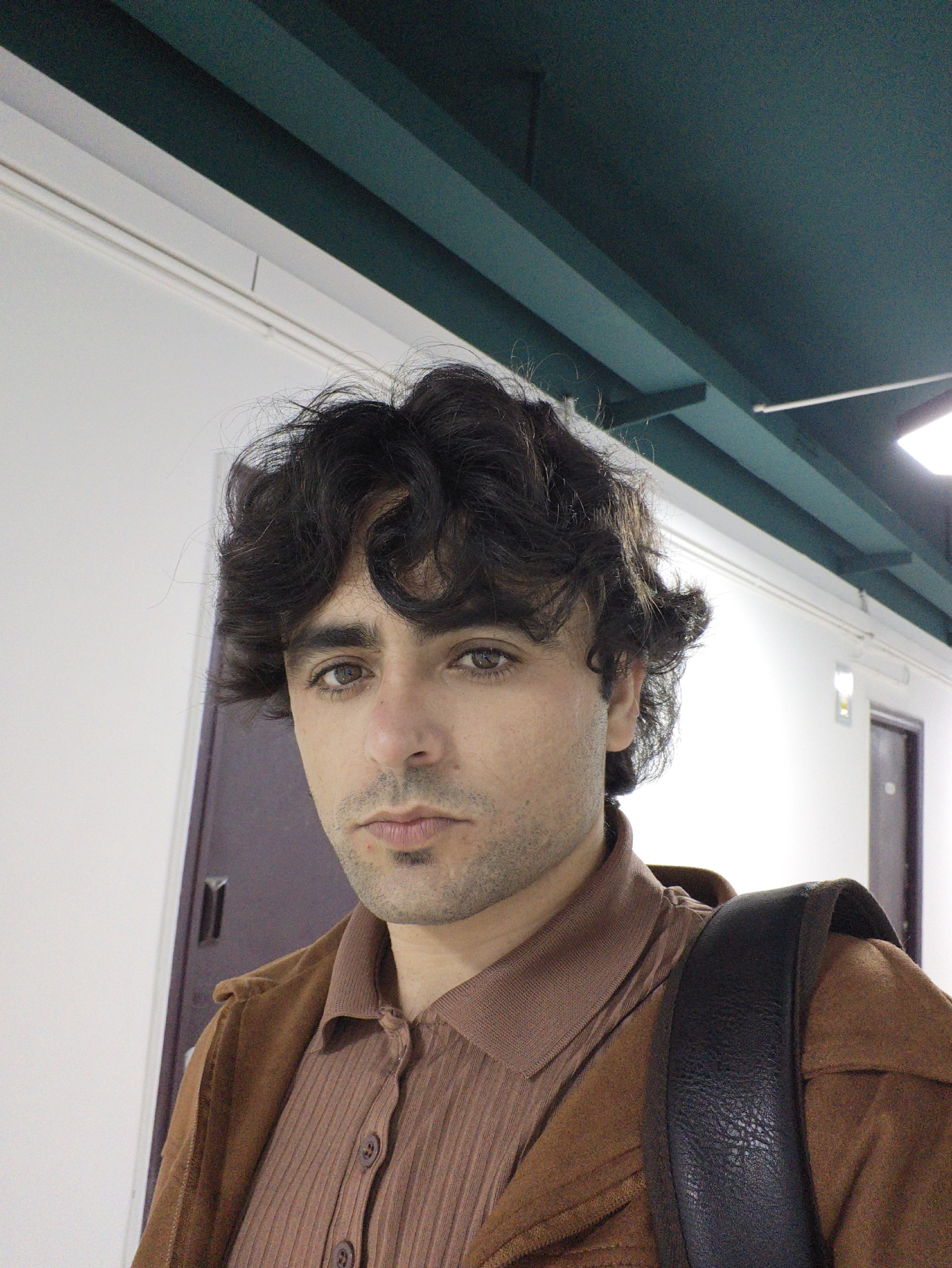 A close-up portrait shot captures an adult male indoors in the Chongqing High-tech Zone, China. The subject, positioned centrally, has dark, curly hair, light brown eyes, and a light stubble on his face. He wears a ribbed, collared brown shirt underneath an open, lighter brown jacket. A black strap, consistent with a backpack, is visible over his right shoulder. The background consists of a plain white wall, above which a dark teal ceiling structure with beams and fluorescent lighting fixtures is visible. Two dark brown doors are partially visible in the background; one to the left of the subject and another further back on the right side. The scene is illuminated by artificial overhead lighting. The subject maintains direct eye contact with the camera.