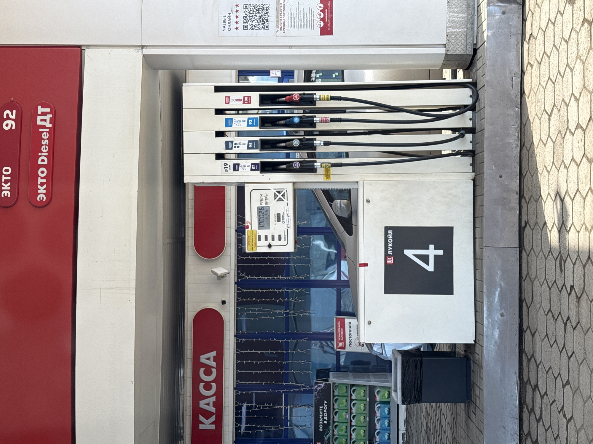 This image shows a gas station, specifically pump number 4, of the LUKOIL brand in Izhevsk, Russia. The pump offers several fuel types, including EKTO 92 and EKTO Diesel. The station operates in a post-payment mode ("ПОСТОПЛАТА"). A sign indicates that gratuities ("ЧАЕВЫЕ") are accepted online. There is a register ("KACCA") and a variety of drinks available ("ВОЗЬМИТЕ В ДОРОГУ") for sale.