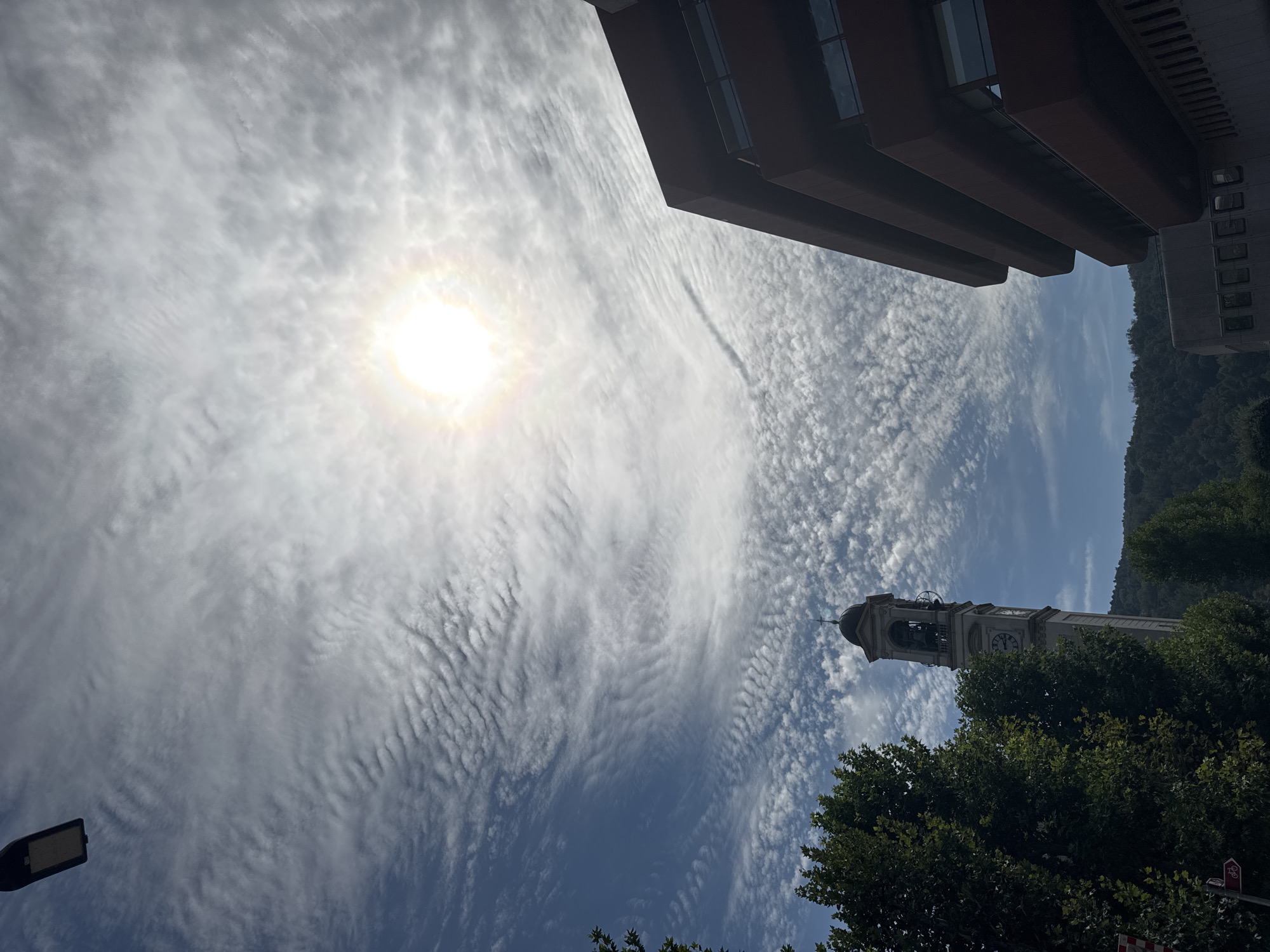 The image depicts an upward-looking view of the sky and some urban structures in Chiasso, Switzerland. In the upper left corner, a dark, rectangular street lamp is visible against a sky filled with wispy, undulating clouds. The sun is prominently displayed in the upper right quadrant, partially obscured by the clouds, creating a bright, halo-like effect.
On the left side of the frame, a tall, slender clock tower with a bell and a dark dome is partially visible behind lush green trees. The clock face is discernible. To the right, a modern, multi-story building with a reddish-brown facade and horizontal balconies is a dominant feature, its angular structure contrasting with the natural elements. The background shows a gentle slope of a tree-covered hill. The scene is captured during daylight.
