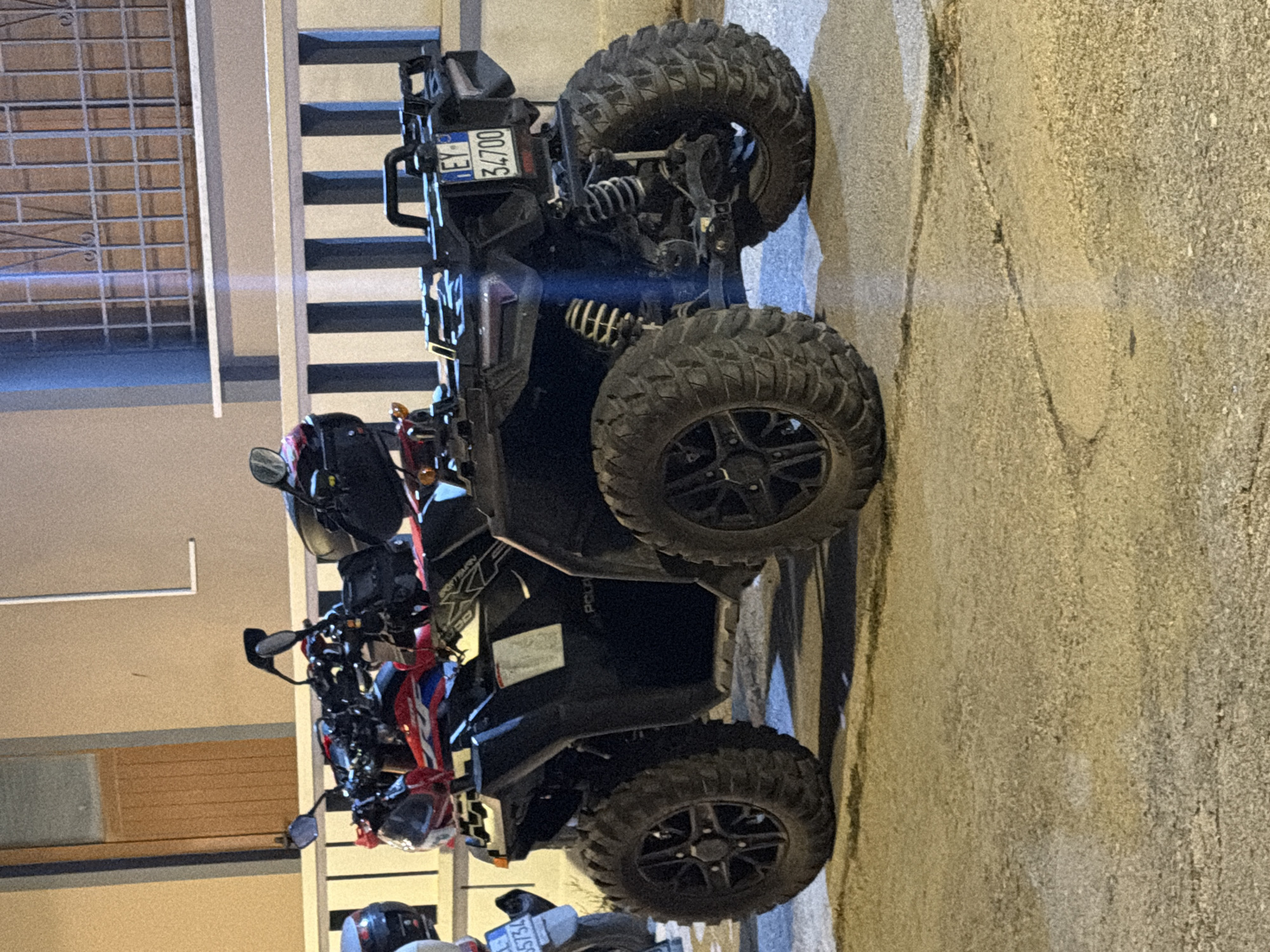 The image shows a black Polaris ATV parked on a paved surface in front of a building with vertical pillars and a barred window. The ATV has large, knobby tires and a red and black body. A license plate with the characters "EY 34700" is visible on the rear of the vehicle. To the left of the ATV, a white motorcycle is partially visible. The lighting suggests it is nighttime. The environment appears to be an urban or suburban setting in the Municipal Unit of Alyzia, Greece, based on the presence of a European-style license plate.