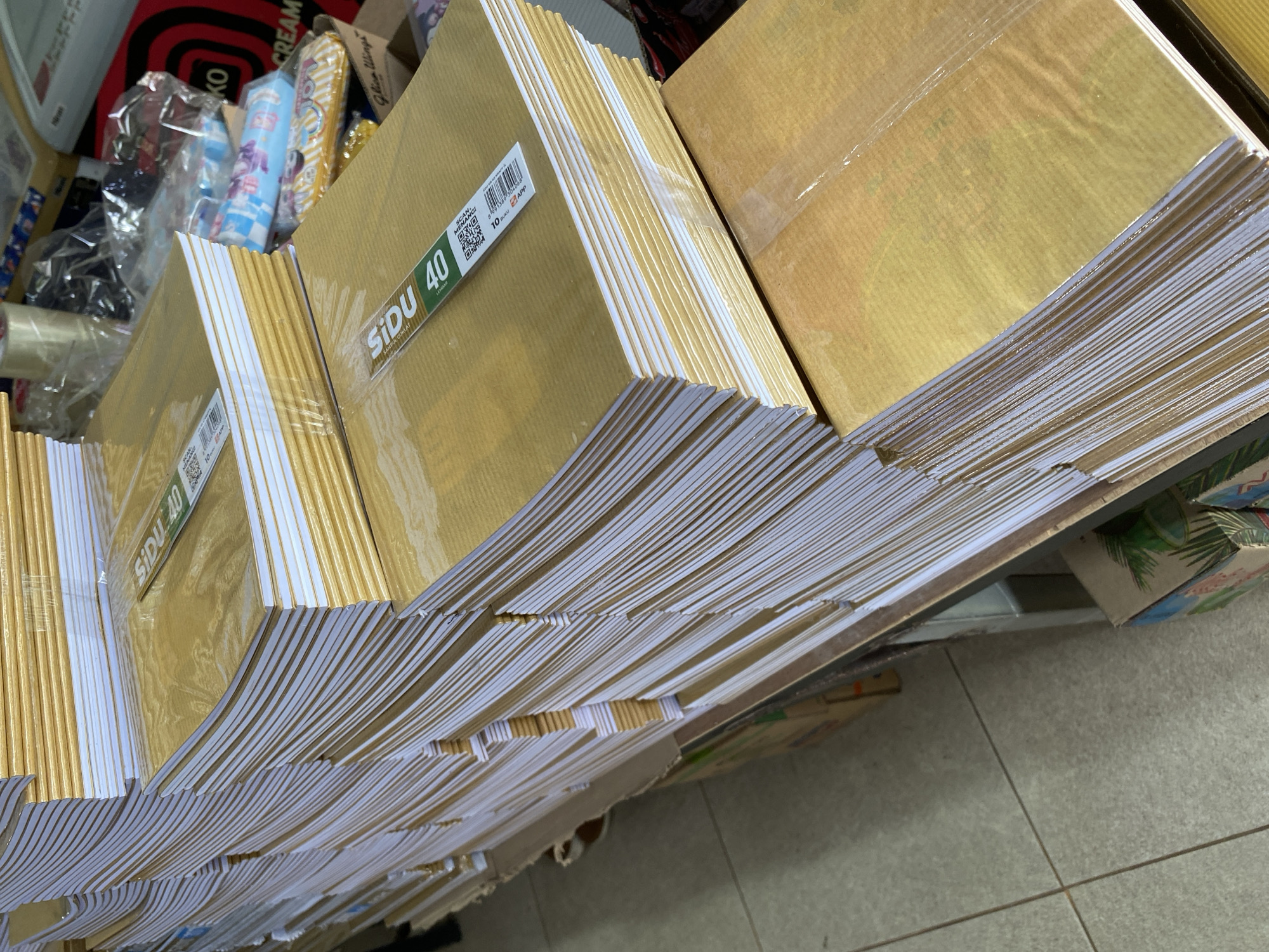 The image depicts a retail store in Tanjong, Indonesia, likely a stationery or general goods shop, with stacks of notebooks and other products. In the foreground, several tall stacks of brown-covered notebooks are arranged on shelves and on the floor. The notebooks are wrapped in plastic, and some have labels indicating "SIDU 40". In the background, there are more shelves filled with various items, including boxes of tissues, packaging tape, and other retail goods. The flooring is tiled, and the lighting suggests an indoor setting. There are no people visible in the frame.